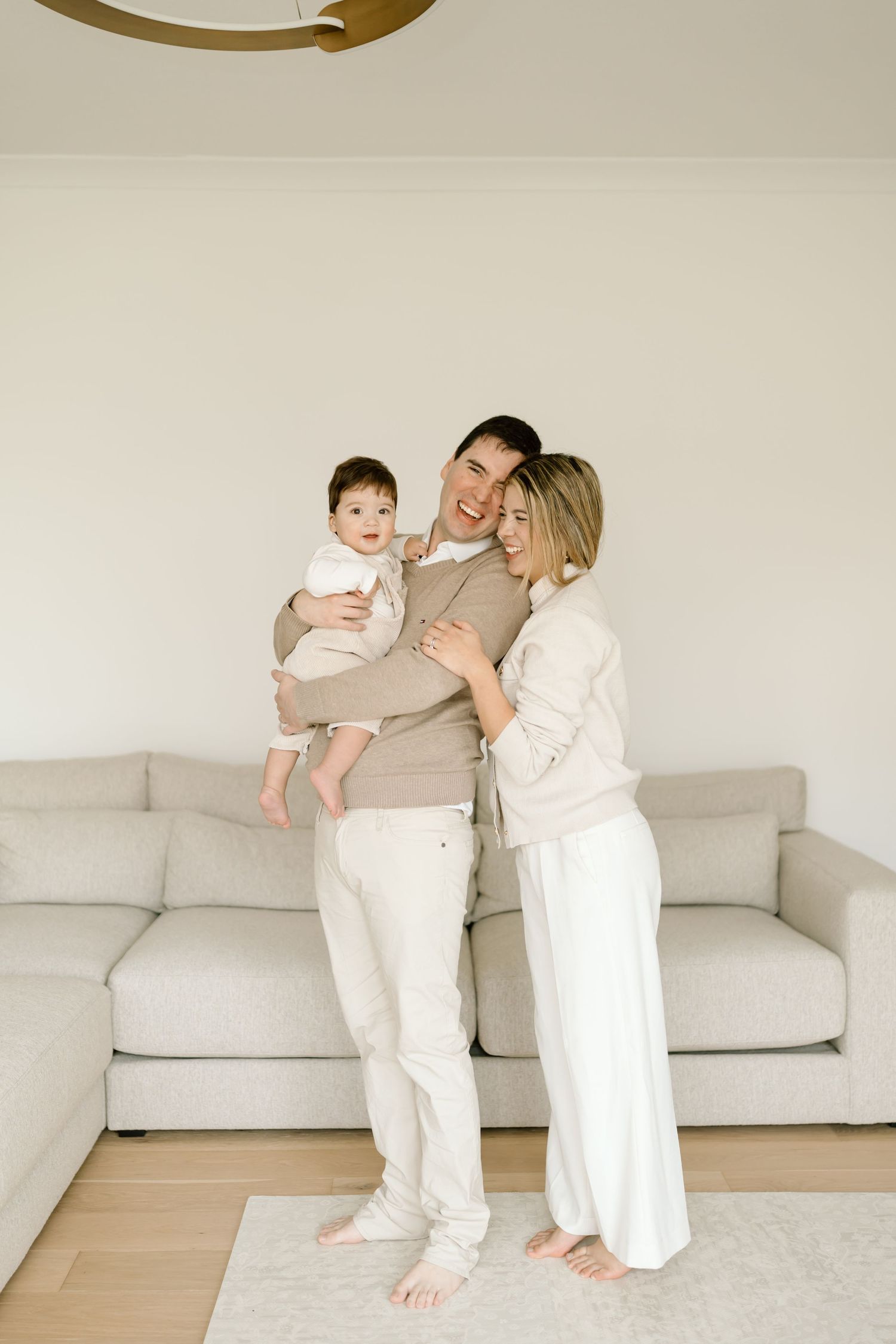 A family in neutral colored clothing stands together on a light gray rug in their modern living room with a beige sofa.