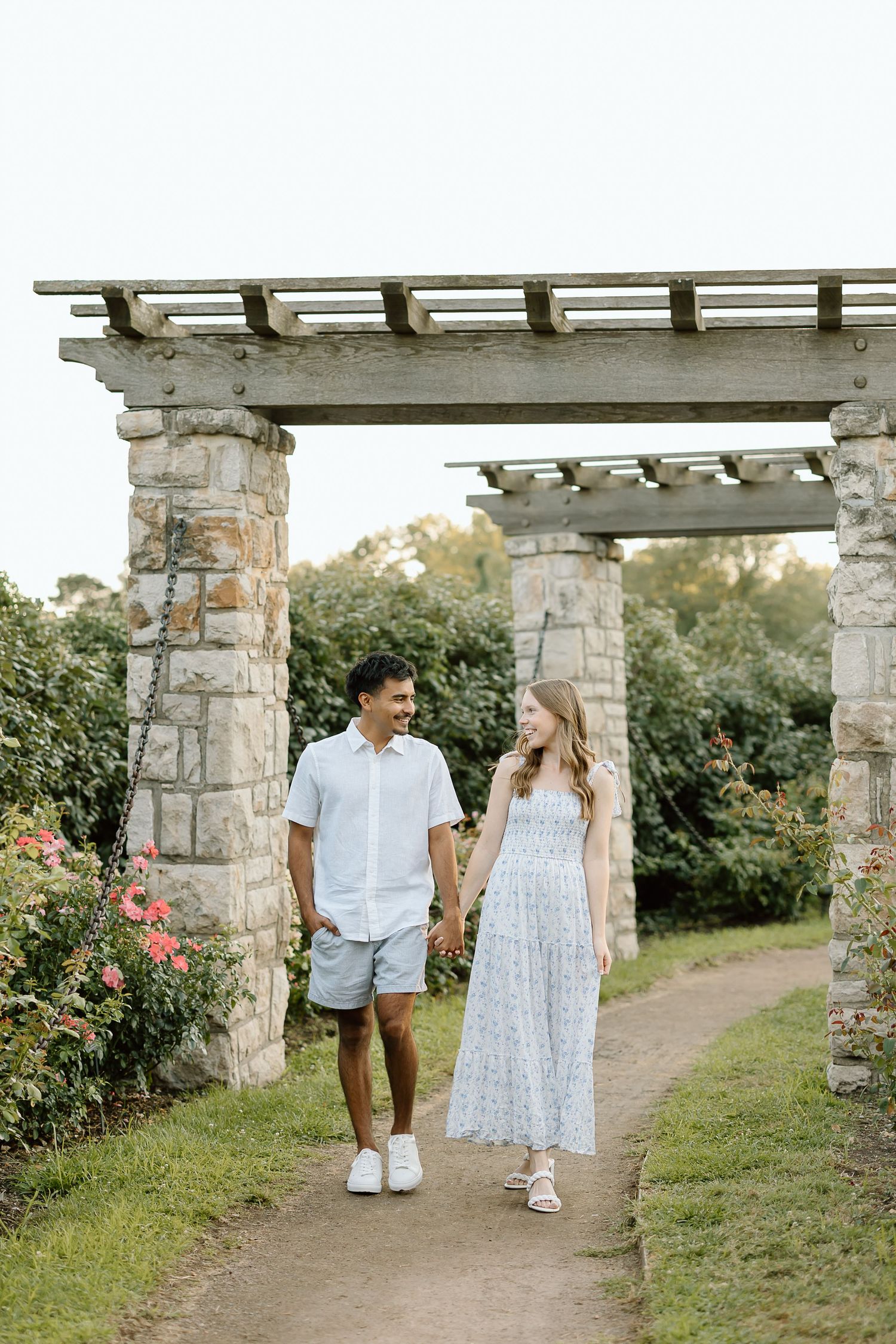 A couple walks together through a romantic garden pergola with stone pillars and blooming roses on a sunny day.