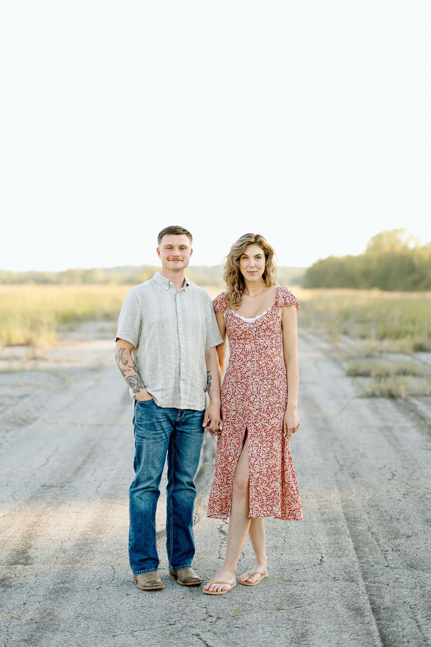A couple stands together on a dirt road at sunset, with her wearing a floral dress and him in casual attire.
