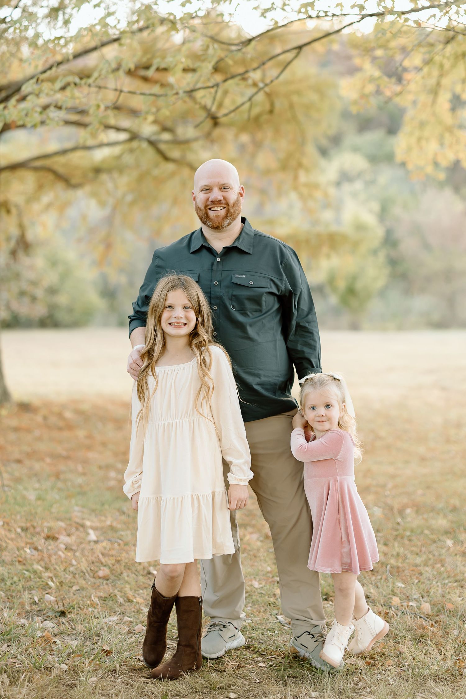 A family wearing coordinated neutral outfits poses together in a scenic autumn field with golden leaves.