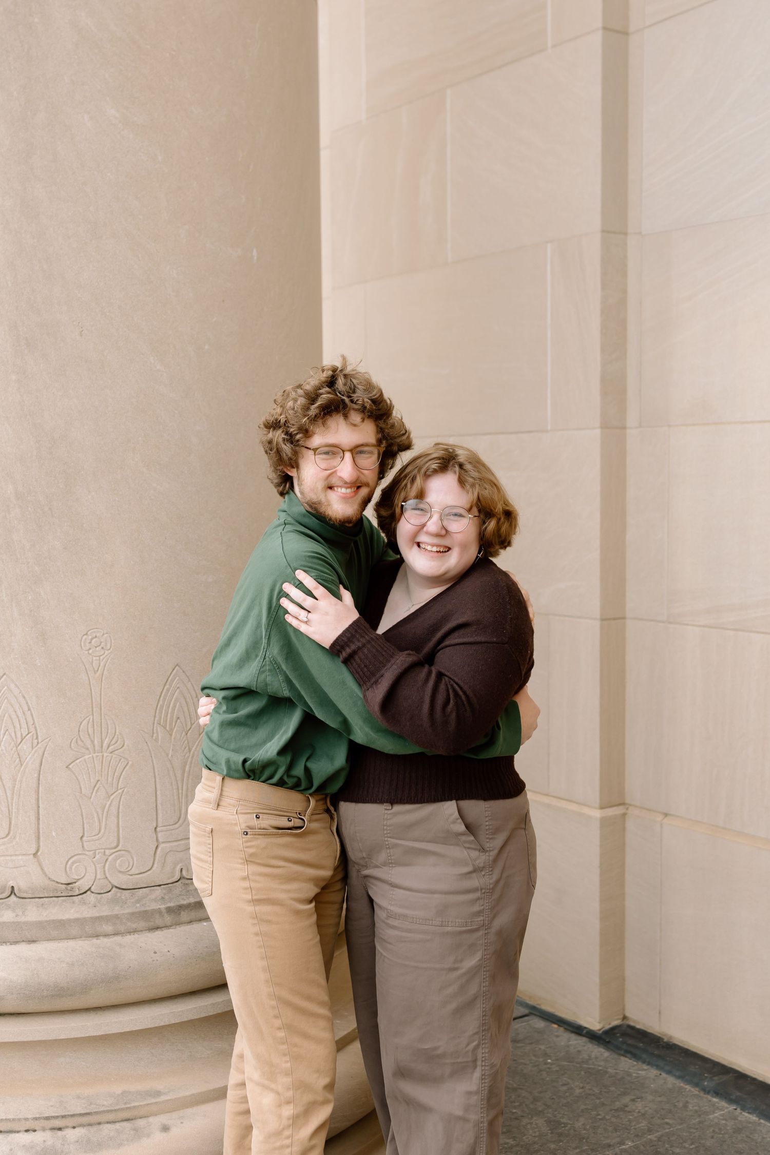 Two people embrace while smiling and standing against a beige stone wall with architectural columns.