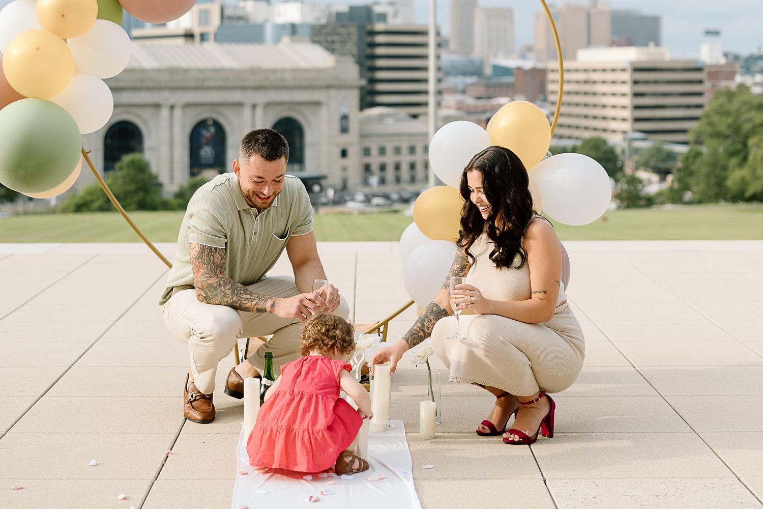 A couple in casual attire kneels on a concrete surface while holding white balloons outdoors on a sunny day.