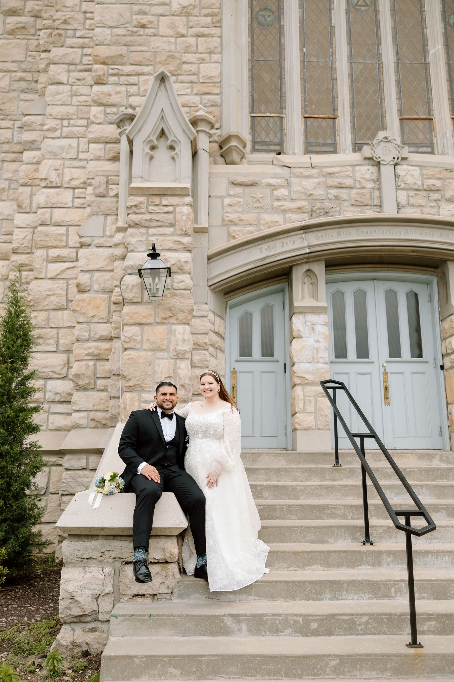 Bride and groom posing on stone church steps, groom in black tuxedo, bride in white lace gown with bouquet.