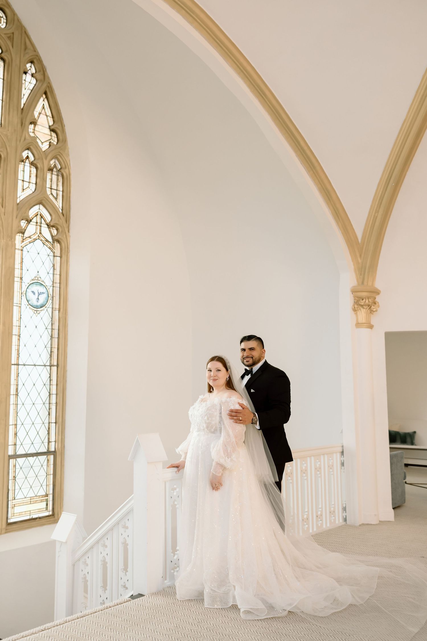 Bride and groom posing elegantly in a white church with gothic arched windows and ornate gold details.
