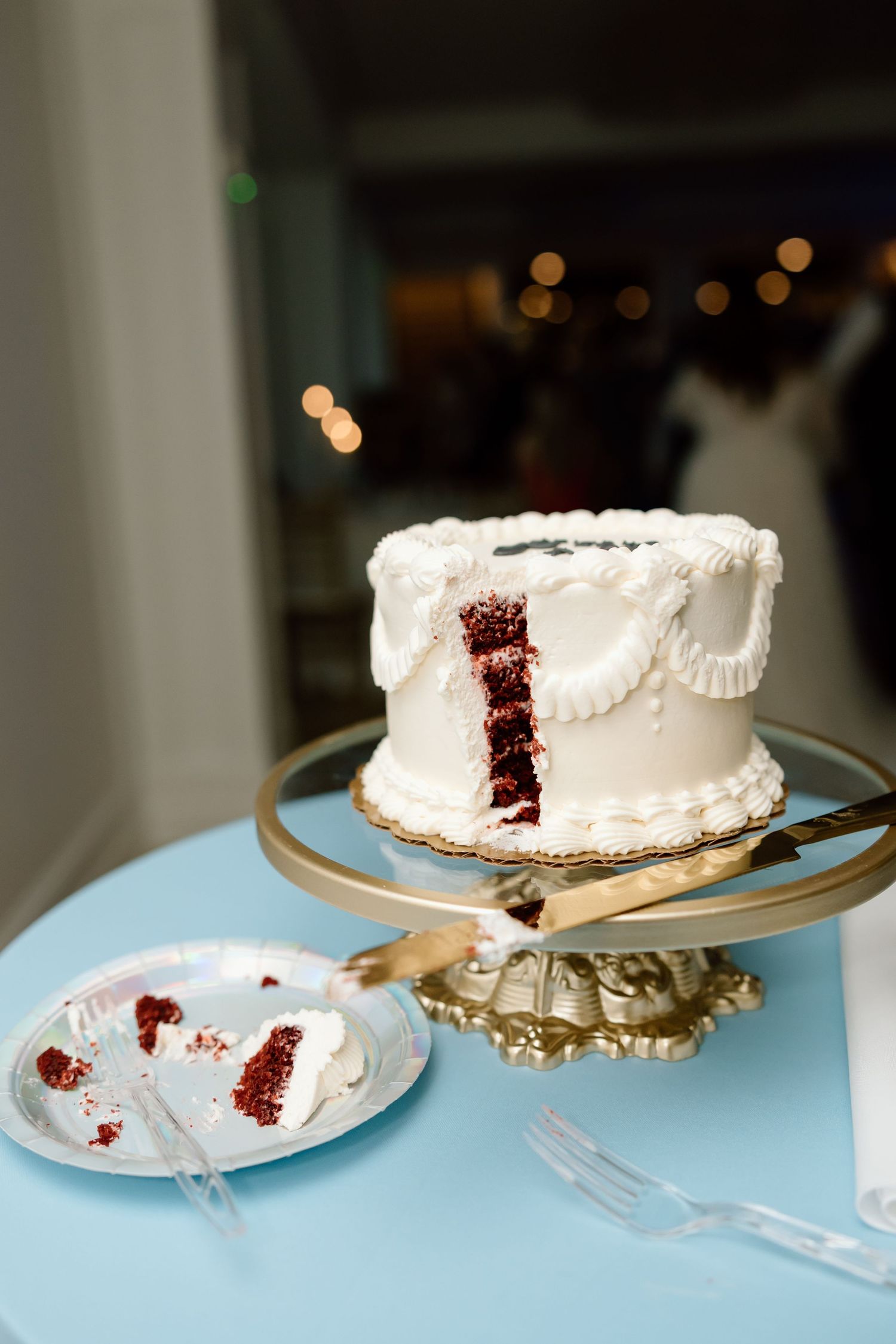 Sliced red velvet wedding cake with white frosting on a gold stand beside a served plate on a blue table.