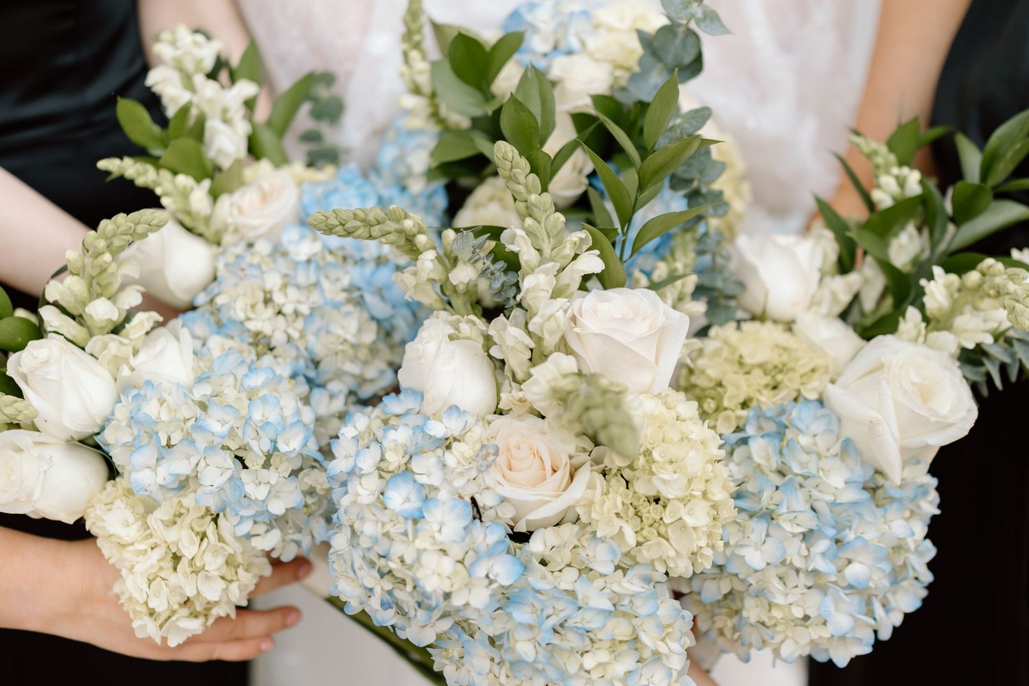 Close-up of elegant bridal bouquets featuring soft blue hydrangeas, white roses, and greenery held by bridesmaids.
