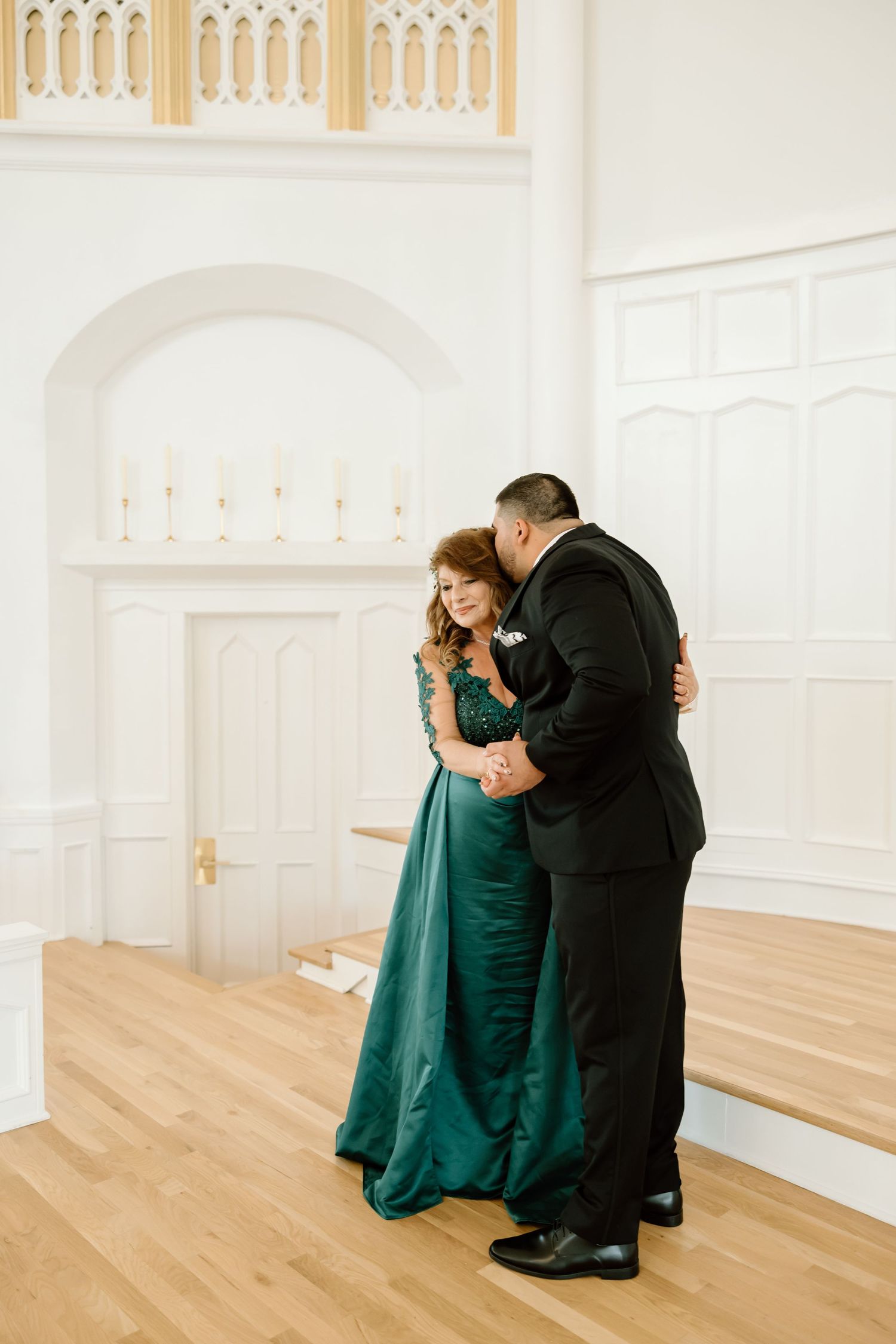 A couple shares a tender dance in an elegant white venue, the woman wearing a stunning emerald green gown.
