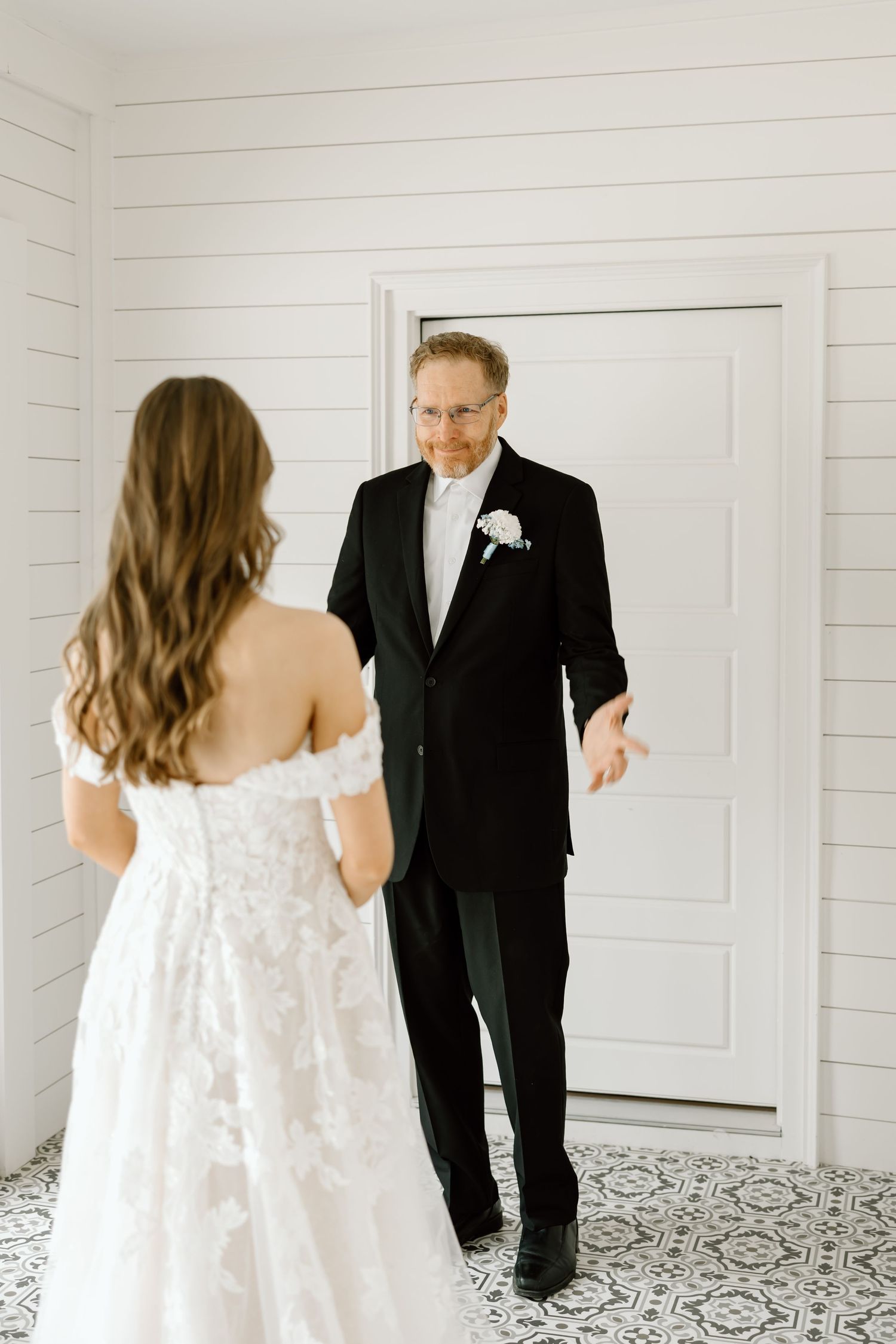 Father in black tuxedo reacts emotionally seeing bride in white lace off-shoulder wedding gown during first look moment.