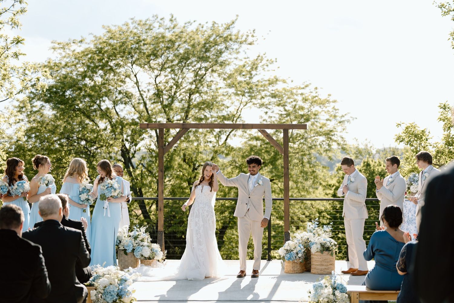Outdoor wedding ceremony with bride and groom at wooden arch, bridesmaids in blue, surrounded by lush greenery.
