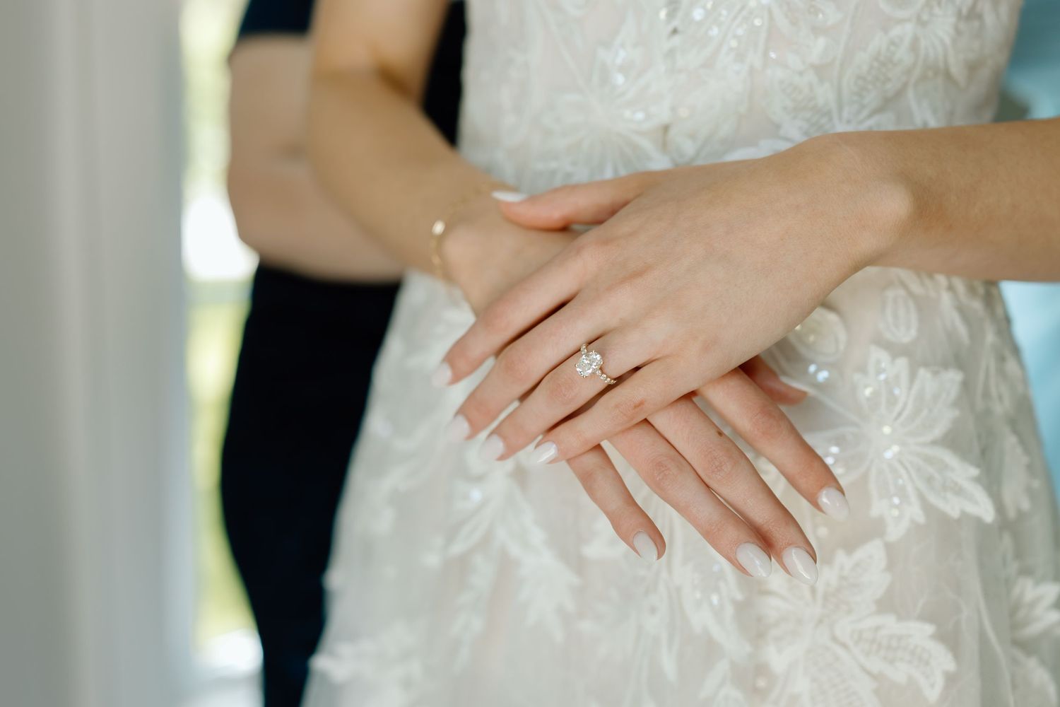 Bride wearing diamond engagement ring with French manicure on white lace wedding dress, groom in background.