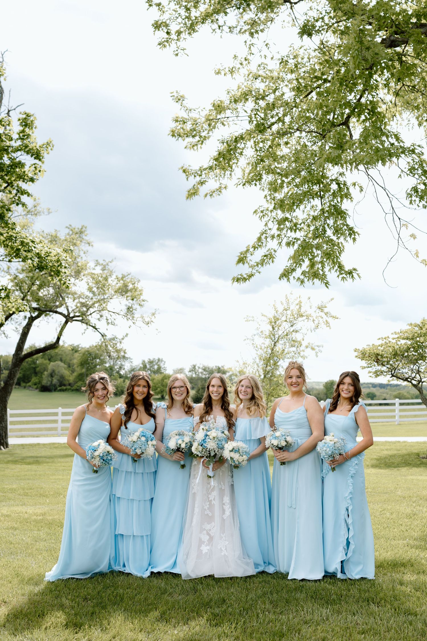 Bride in white lace gown poses with six bridesmaids in light blue dresses holding bouquets outdoors.