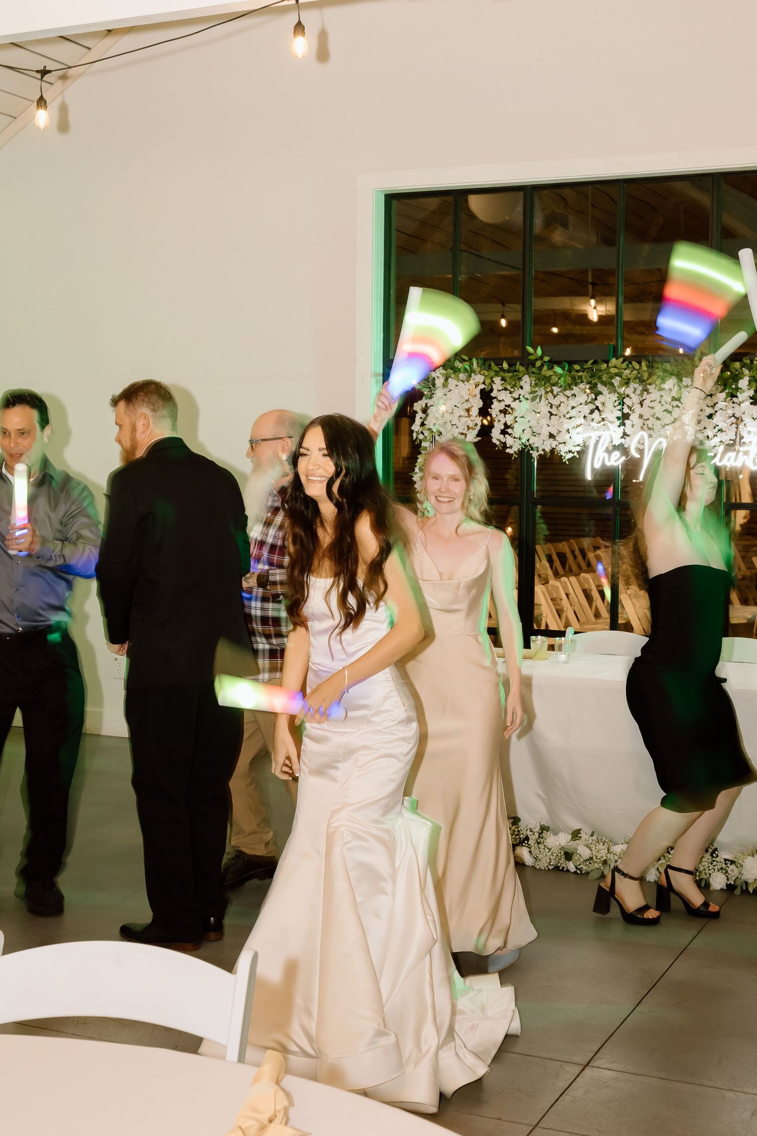Wedding guests dance together at an indoor reception venue with hanging lights and white decor.