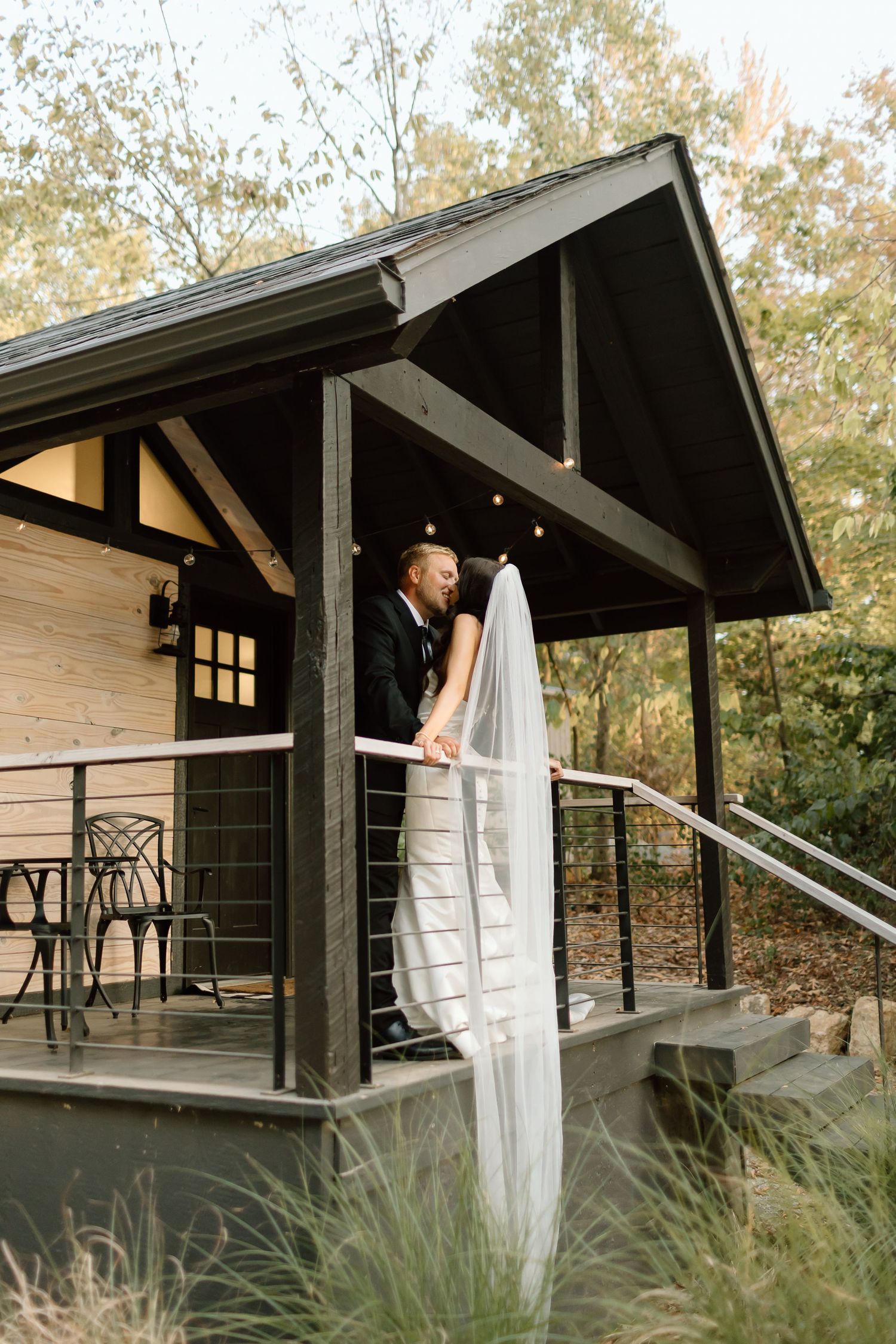 A rustic cabin porch with dark wooden beams and ornate metal railings surrounded by autumn trees and tall grasses.