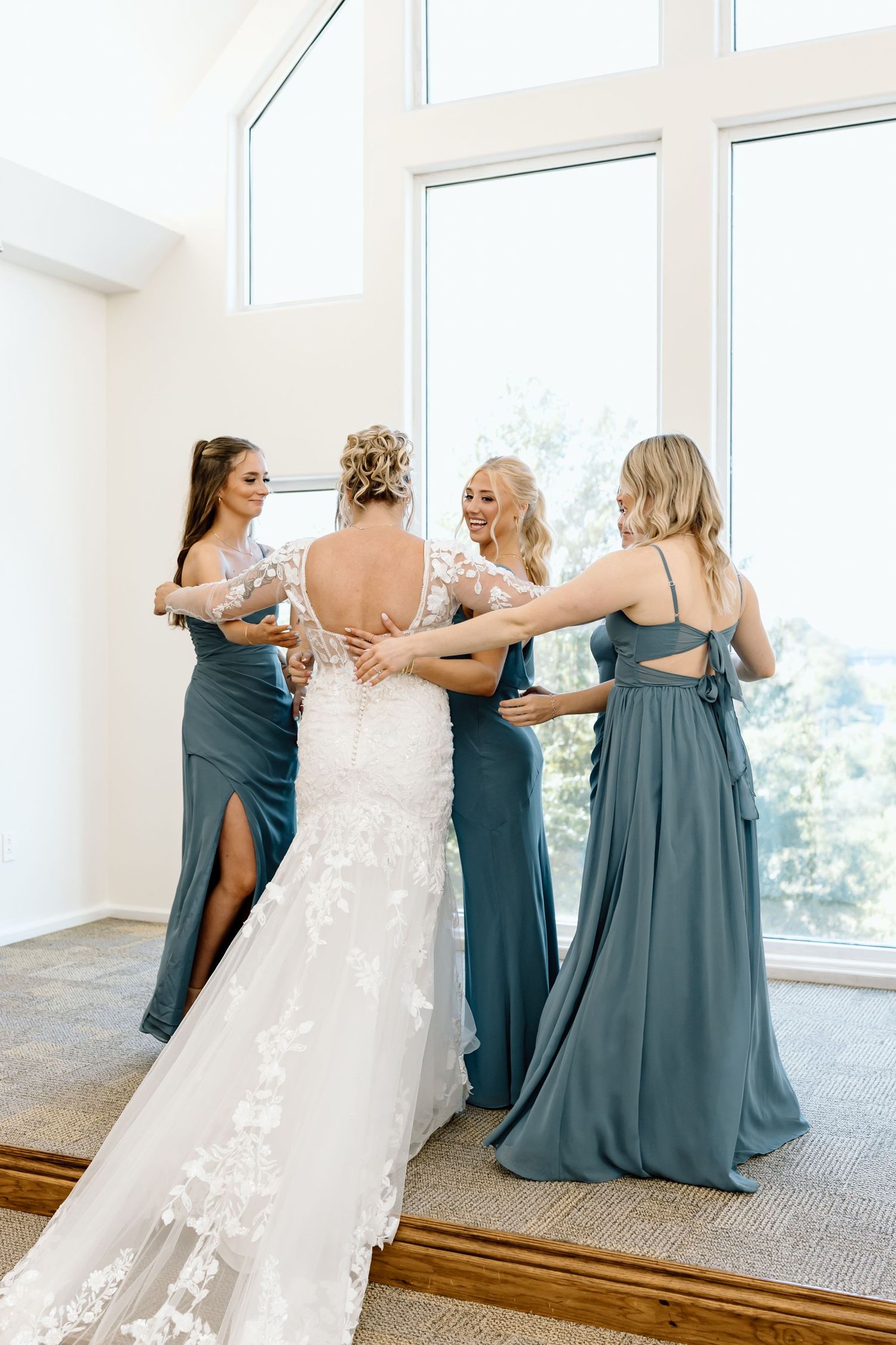 Bridal party in matching teal bridesmaid dresses help adjust the lace train of a wedding gown by large windows.
