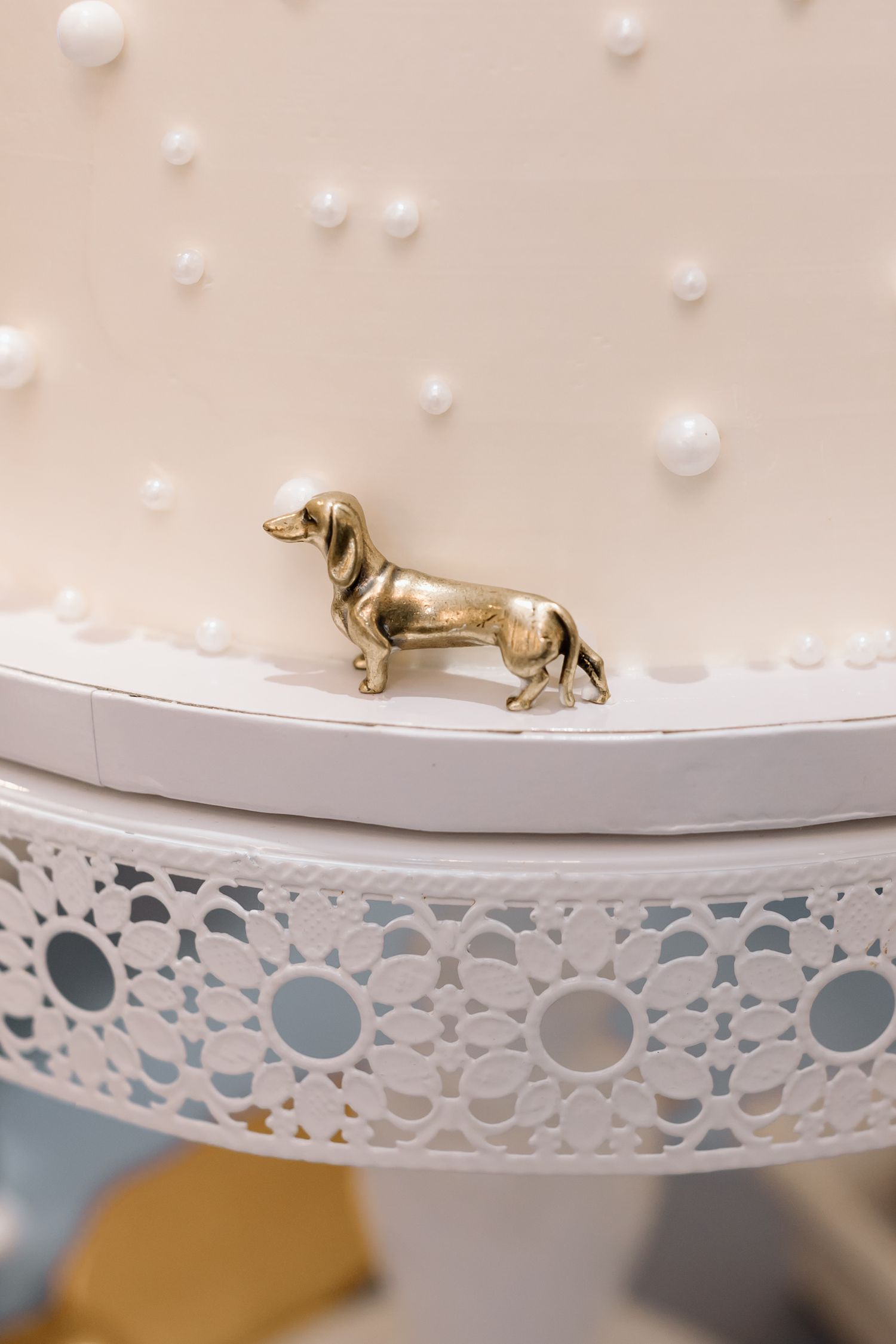 Small golden dachshund figurine sits atop white decorative shelf with delicate lace-like pattern and pearl embellishments.