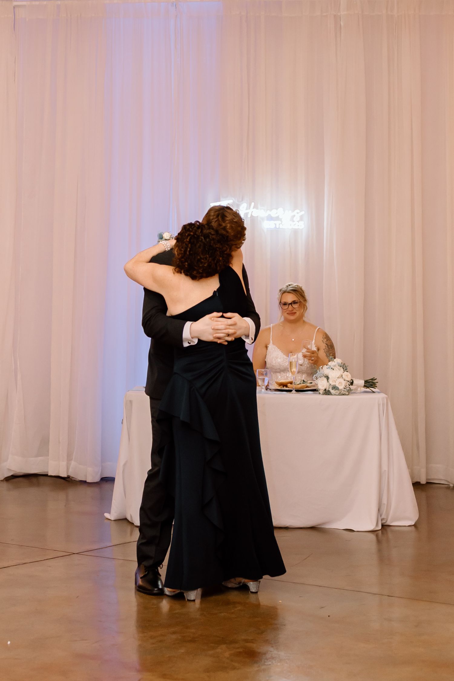 A couple shares a slow dance at a wedding reception while someone watches from a decorated table in the background.