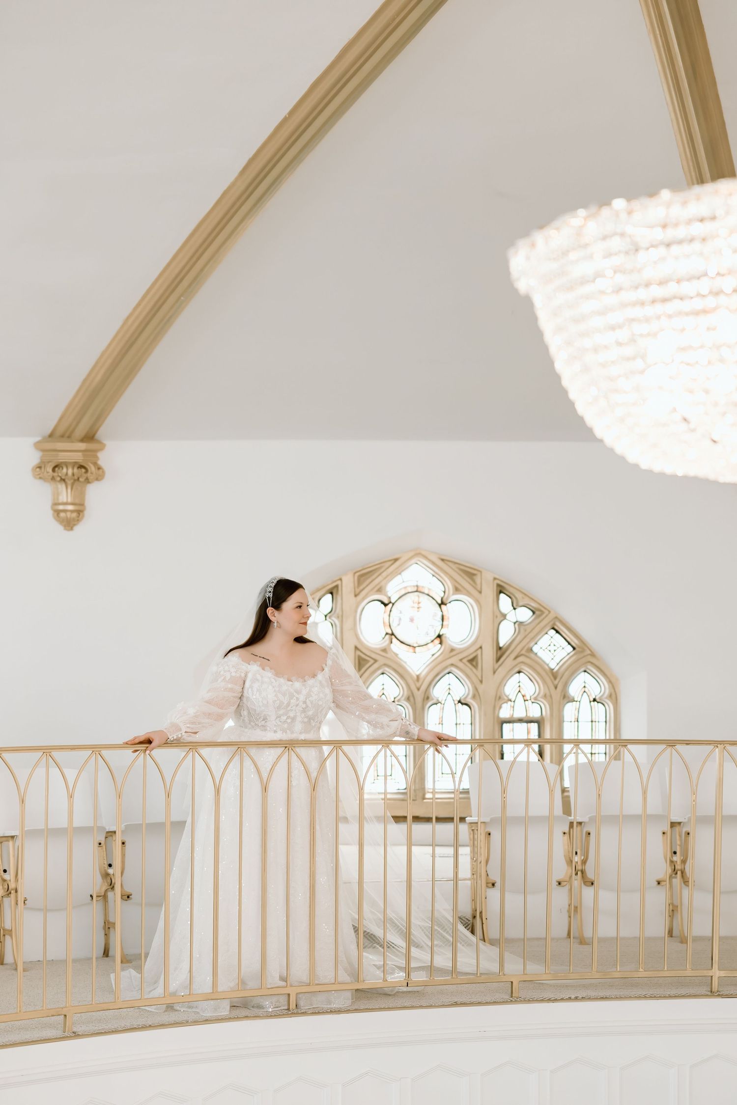Bride in white gown stands on golden balcony railing inside elegant white chapel with Gothic arched windows and chandelier.