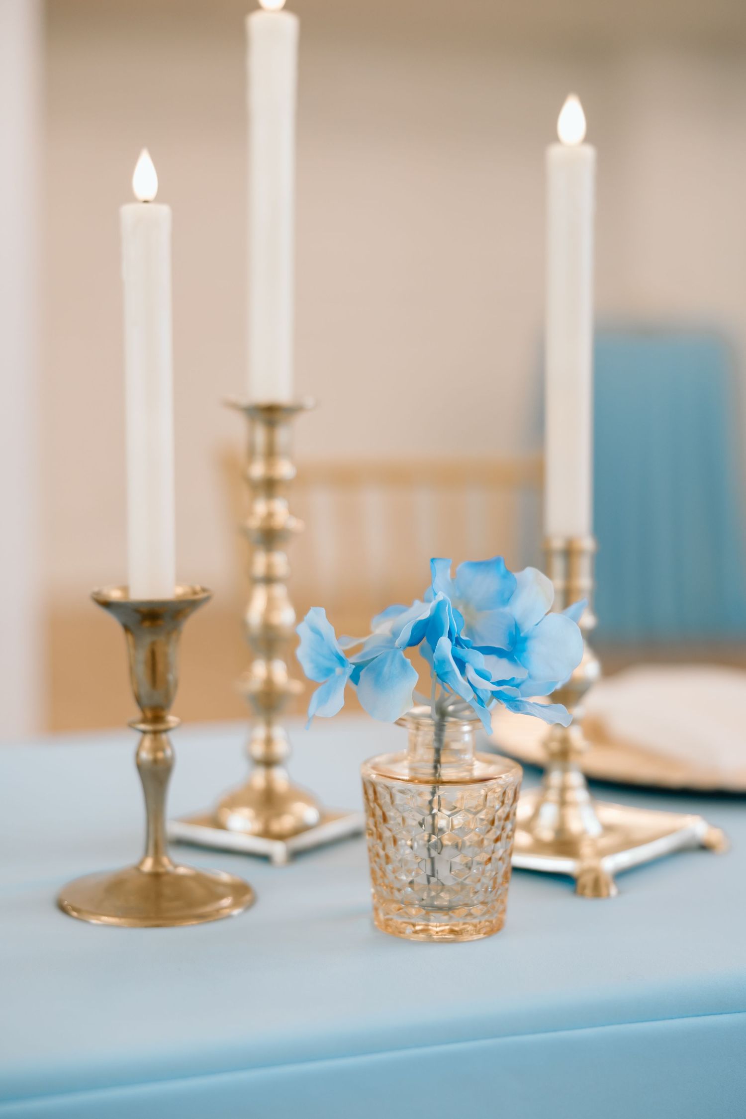 Gold candlesticks with white candles and a crystal vase with blue hydrangeas on a light blue tablecloth.