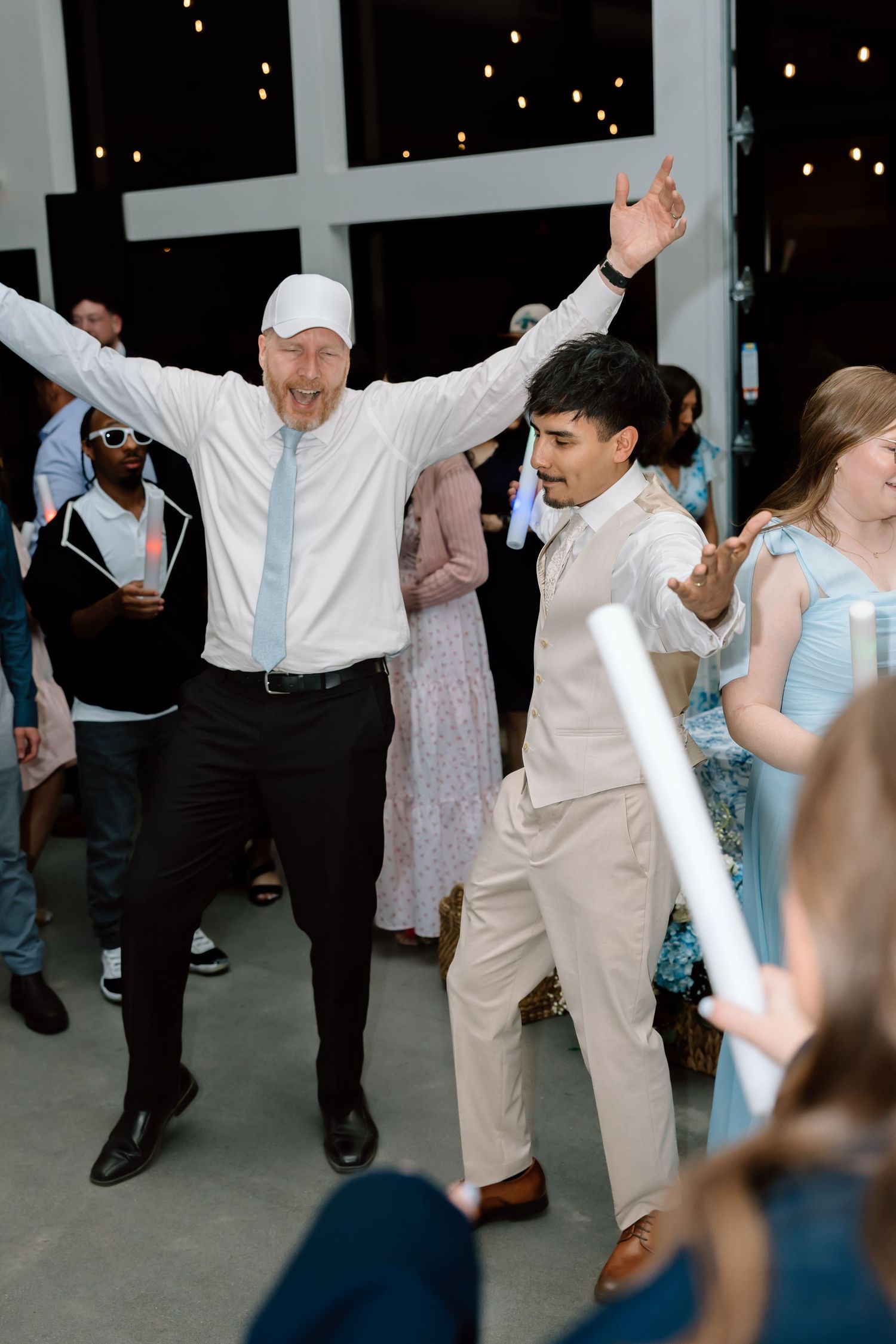 Two men dancing joyfully at a wedding reception, one with arms raised high, surrounded by guests with glow sticks.