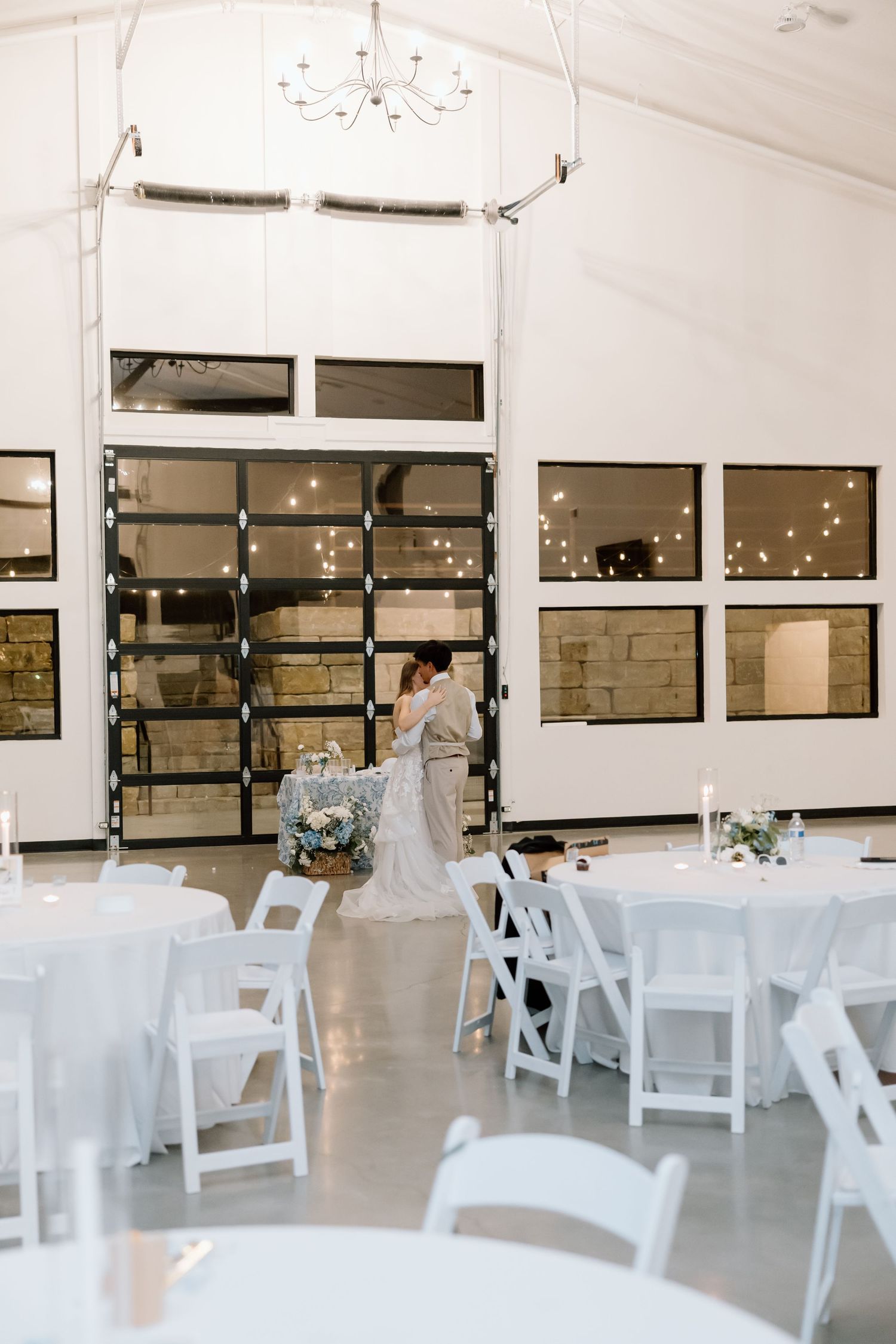 Bride and groom share first dance in elegant white wedding reception hall with chandelier and string lights.