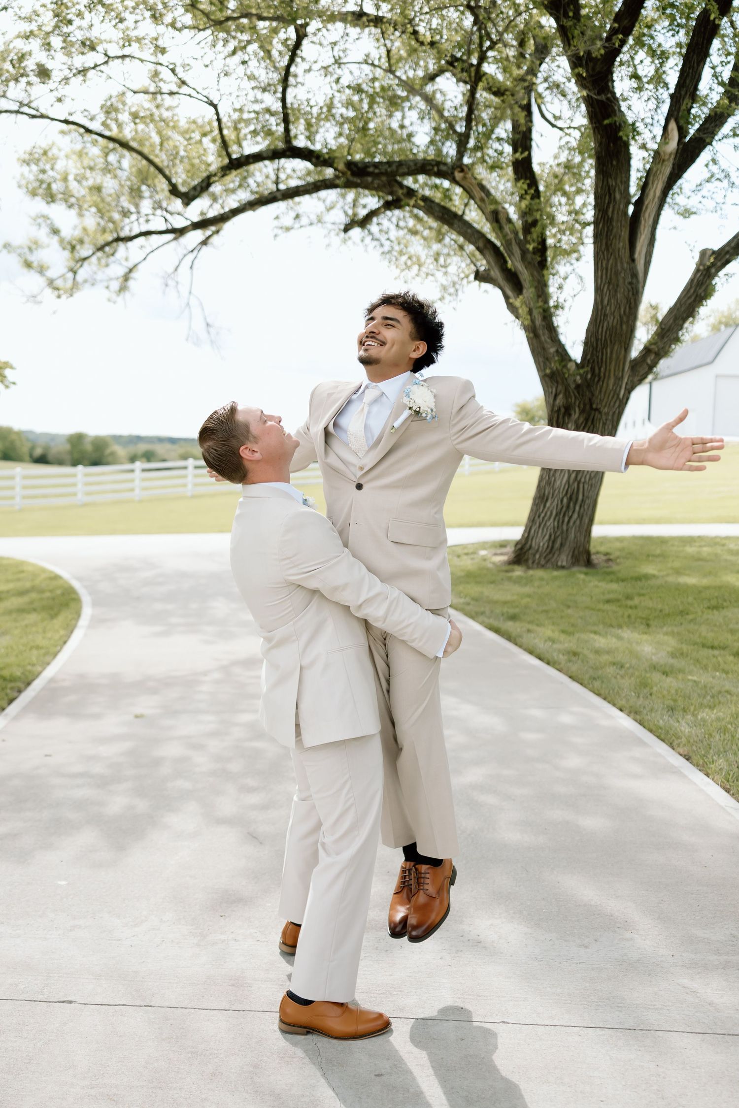 Two grooms in matching beige suits celebrate their wedding day outdoors, one lifting the other joyfully on a scenic pathway.