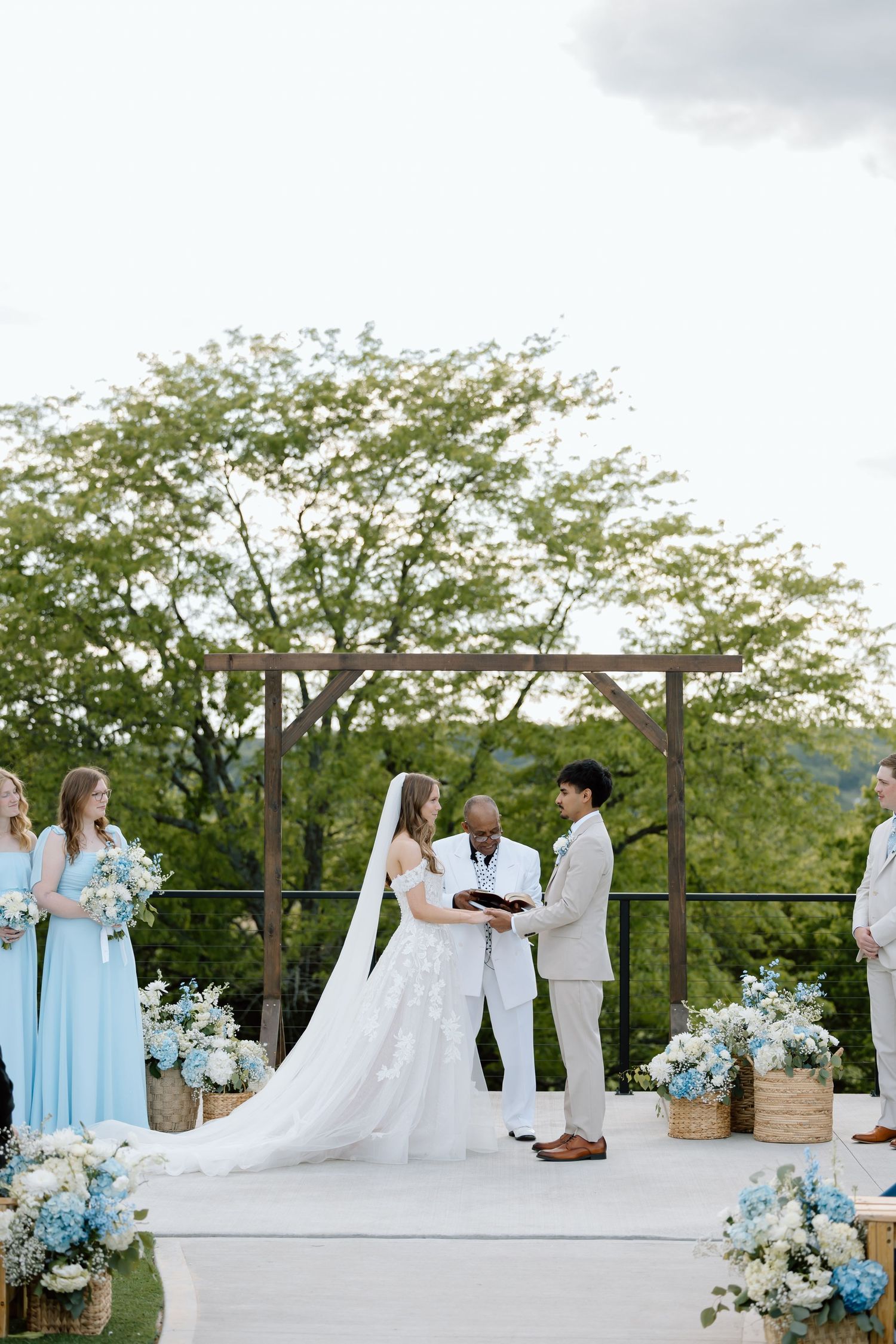Outdoor wedding ceremony with bride in white gown and groom in tan suit exchanging vows under wooden arch with blue floral arrangements.