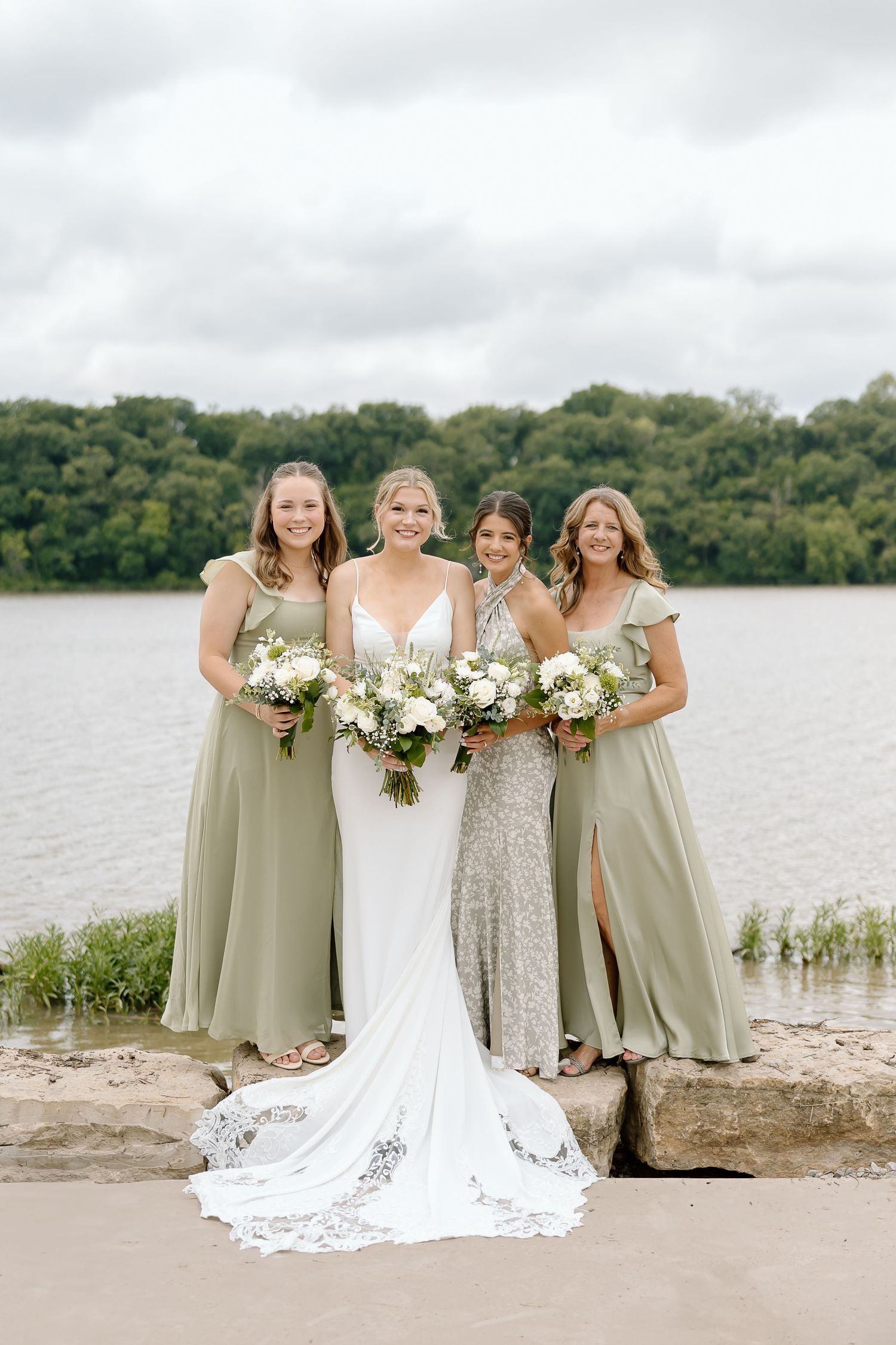 Bridal party in sage green dresses and white gown pose together by a serene lakeside on overcast day.