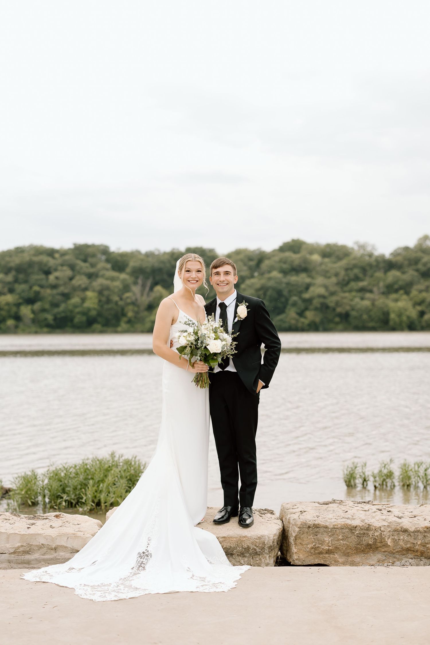 A couple shares an intimate moment by a waterfront in wedding attire with a beautiful lake and greenery in the background.