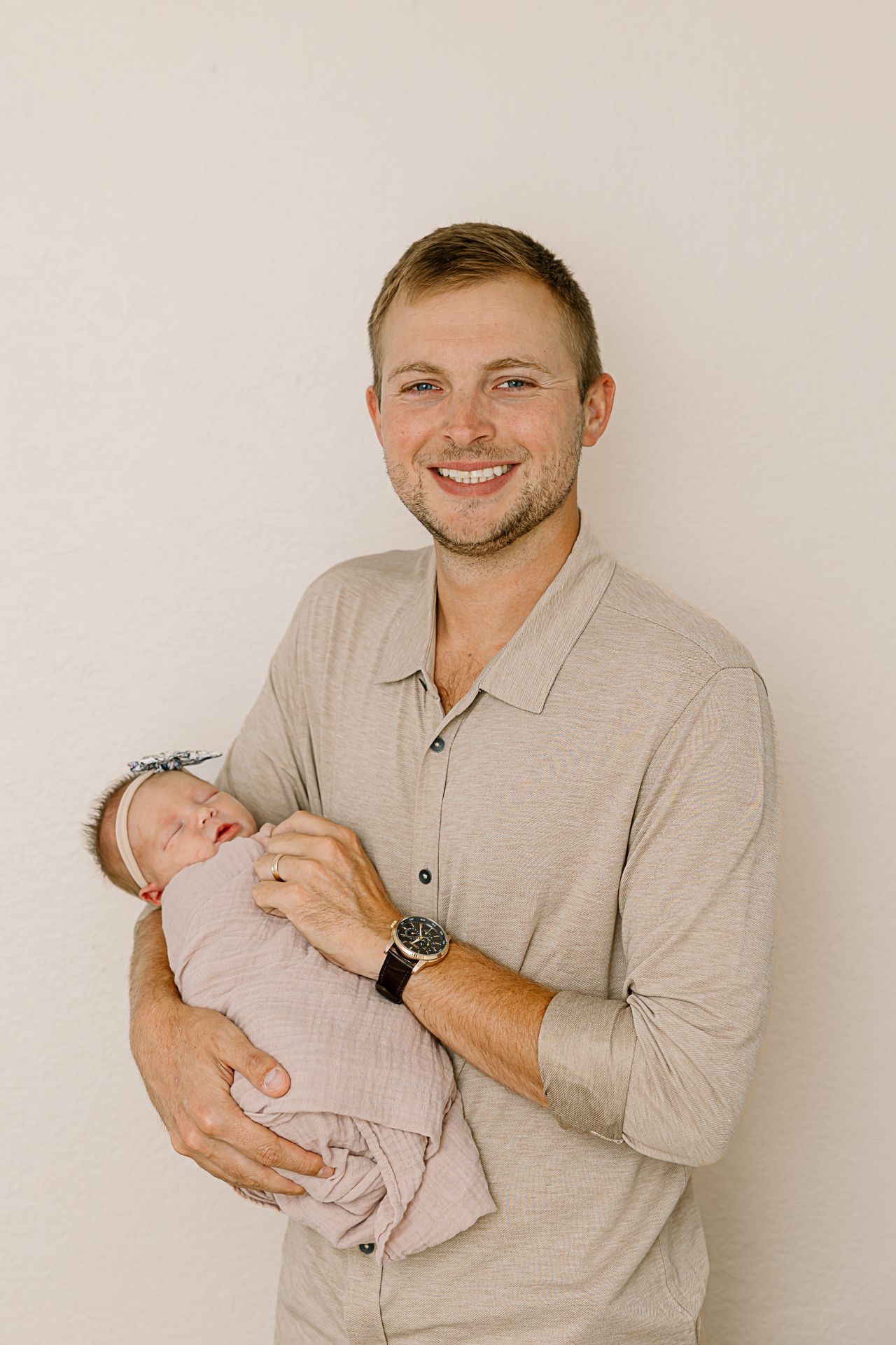 Adult in grey button-down shirt holding sleeping newborn against neutral background.