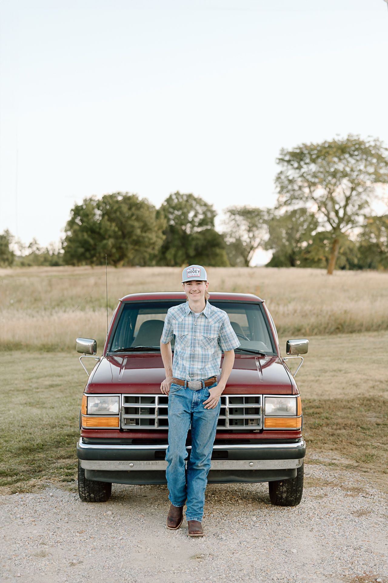 Someone in casual attire standing in front of a classic red Ford pickup truck in a rural setting.