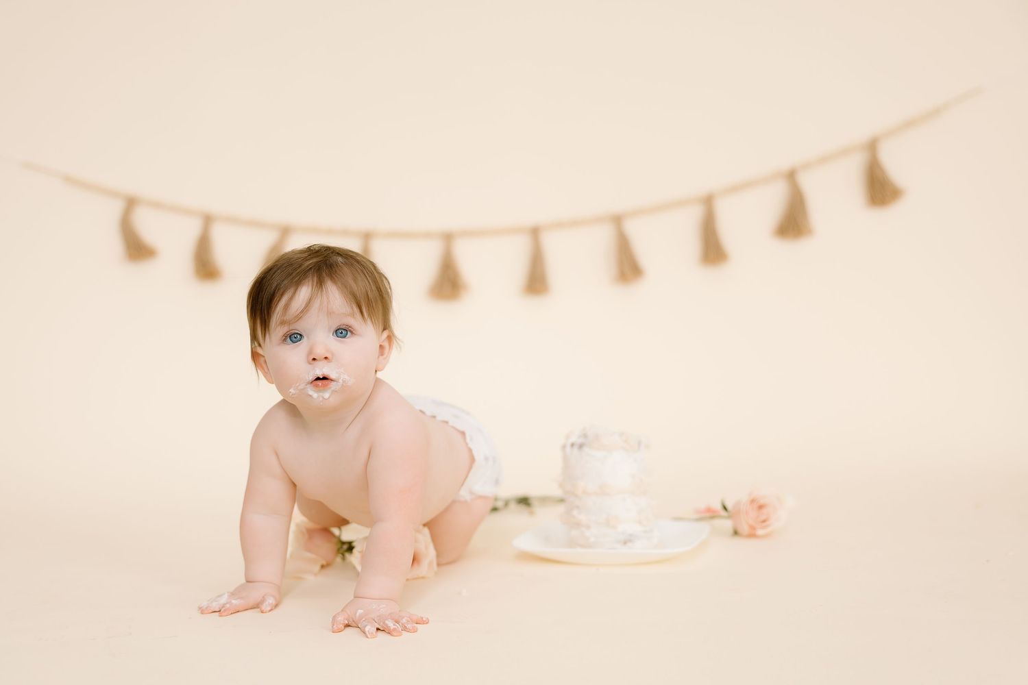 Baby crawling near white cake with tassel garland hanging on beige wall.