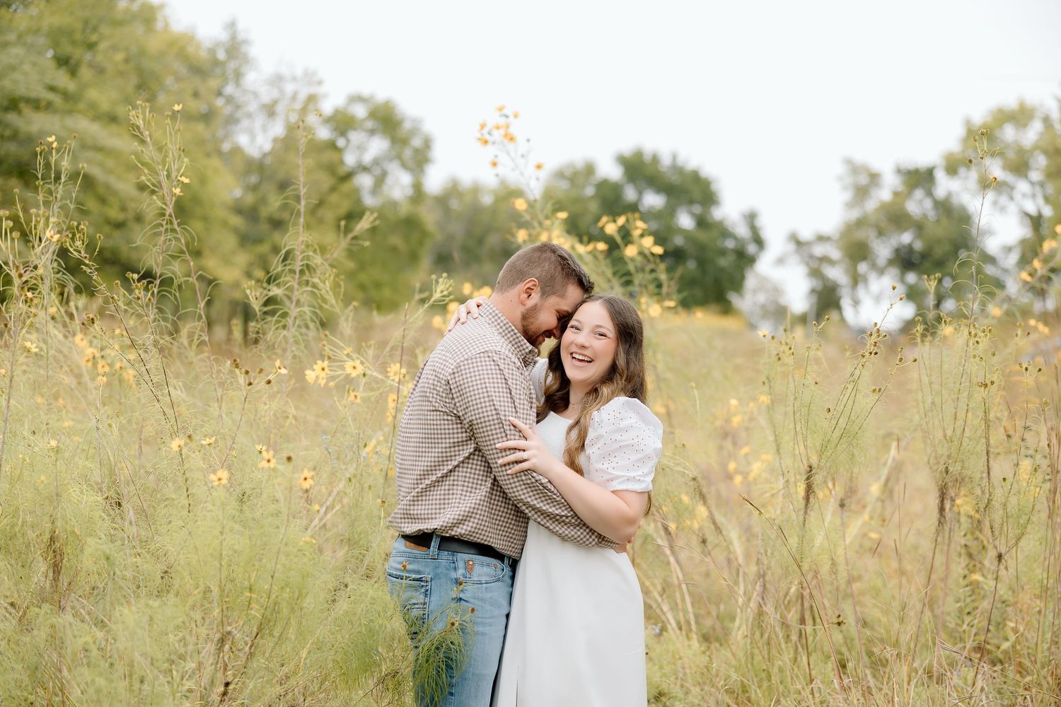 A romantic couple embrace in a sunlit field with tall grass and yellow wildflowers in the background.