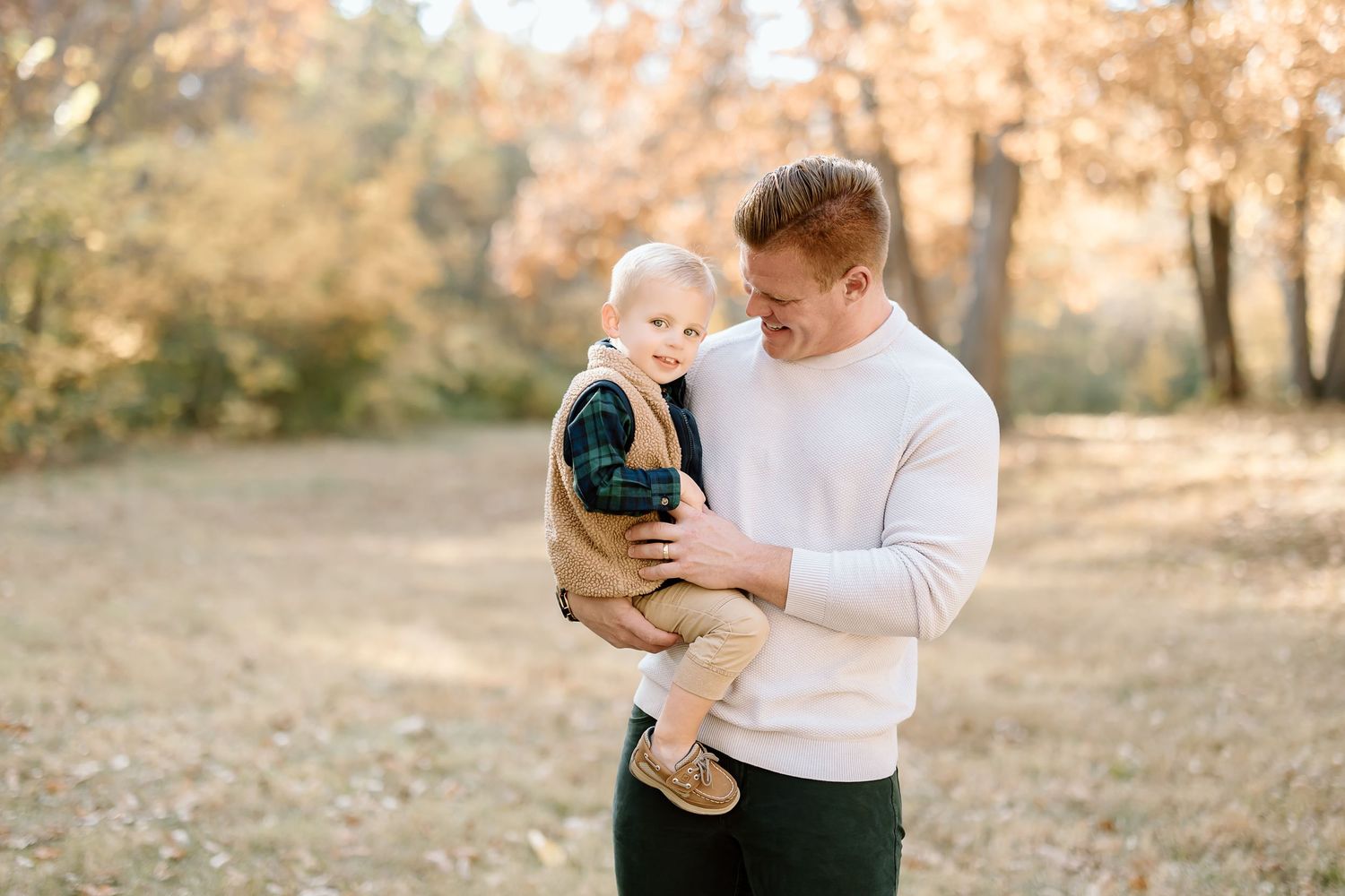 A parent holds a small child wearing a vest in an autumn outdoor setting with golden foliage in the background.