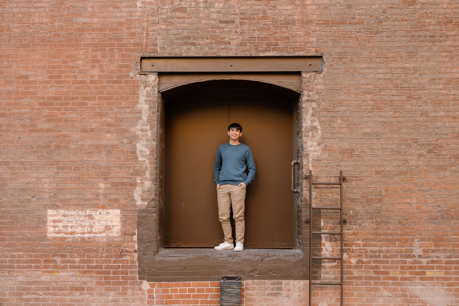Standing in brick doorway wearing casual blue sweater and khaki pants.
