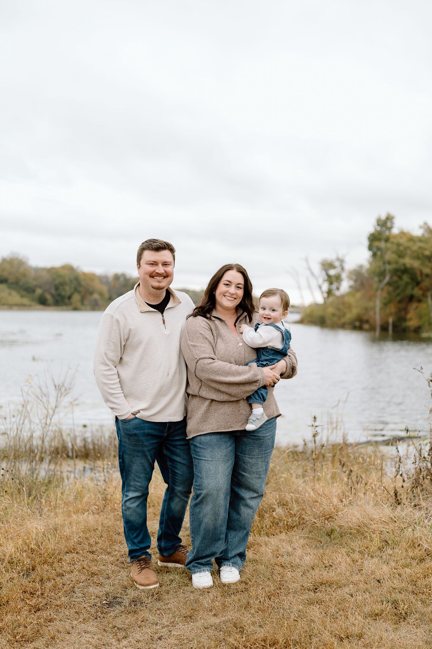 Family of three wearing matching beige sweaters and jeans pose together by a lake on an autumn day.