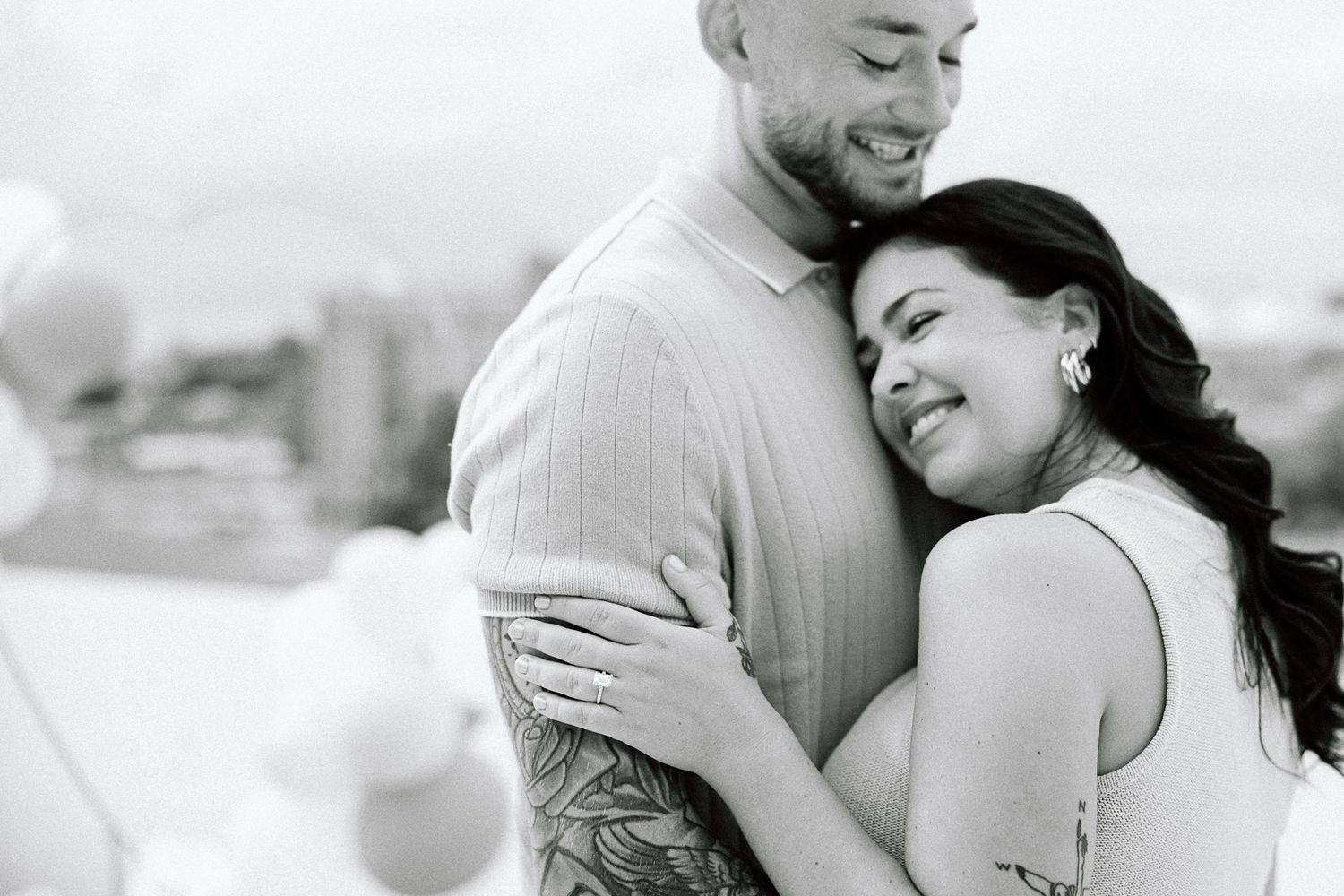 A black and white portrait captures a tender embrace between a couple against an urban backdrop.