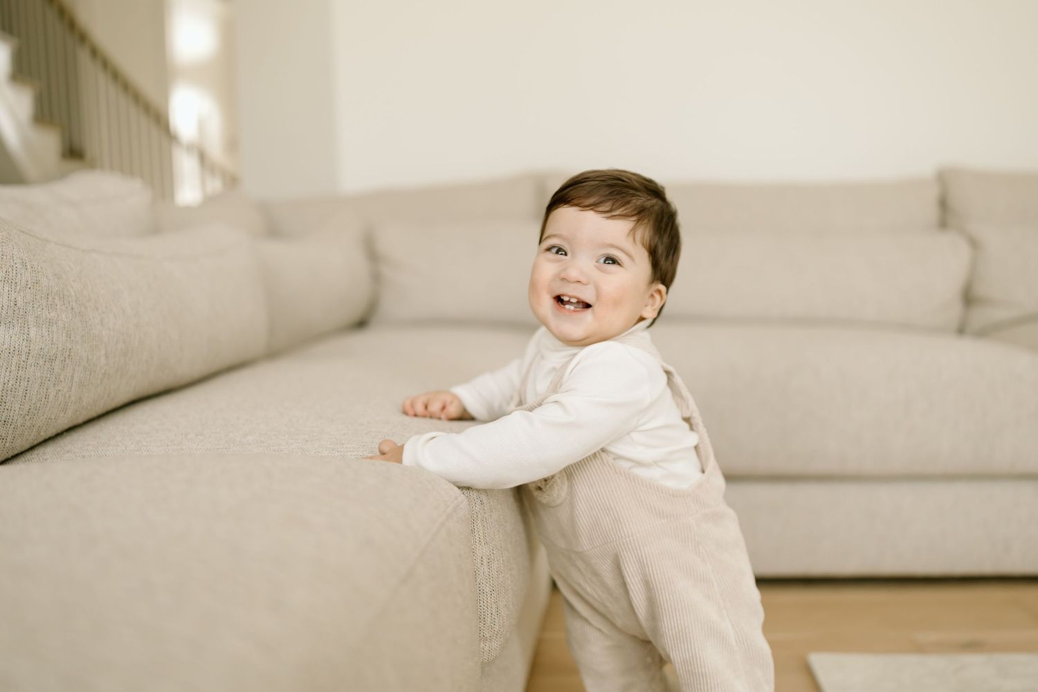 A baby in a white sweater laughs while playing and crawling near a light gray couch.