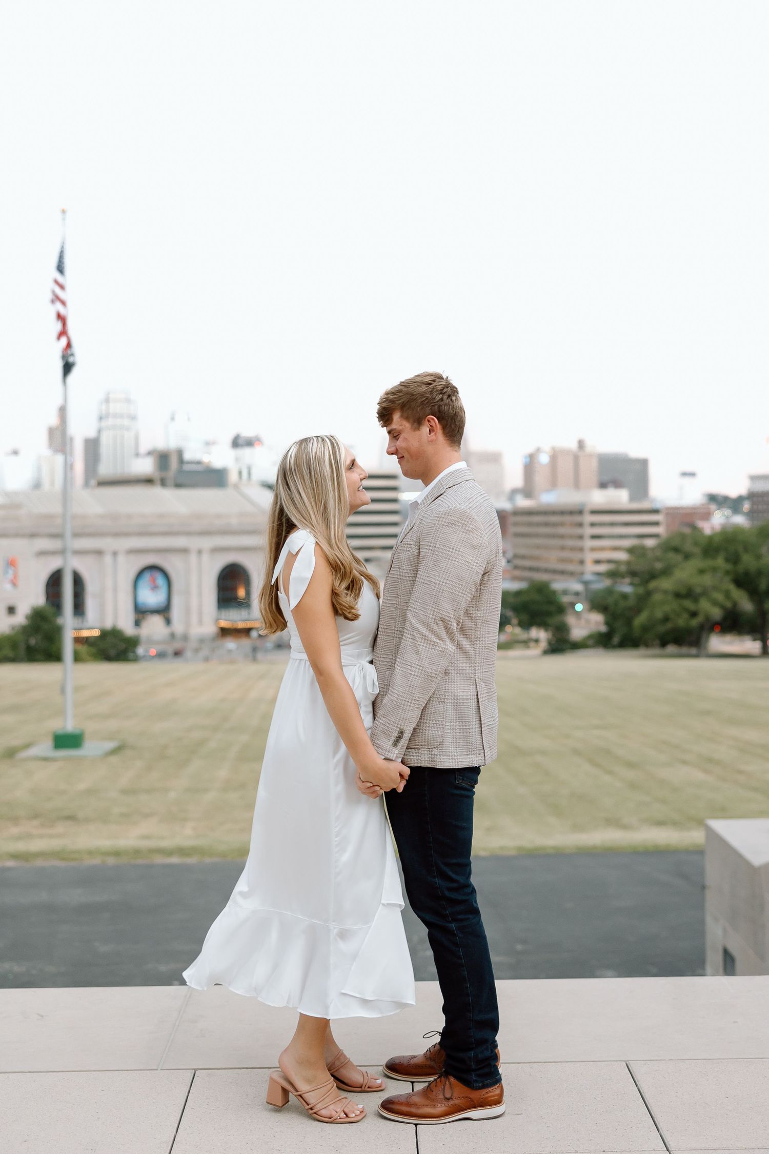 A couple stands close together on a rooftop terrace with an American flag and city buildings in the background.