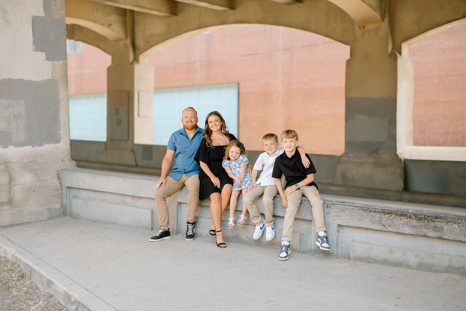 Family sits together on concrete steps outside a brick building for casual outdoor portraits.