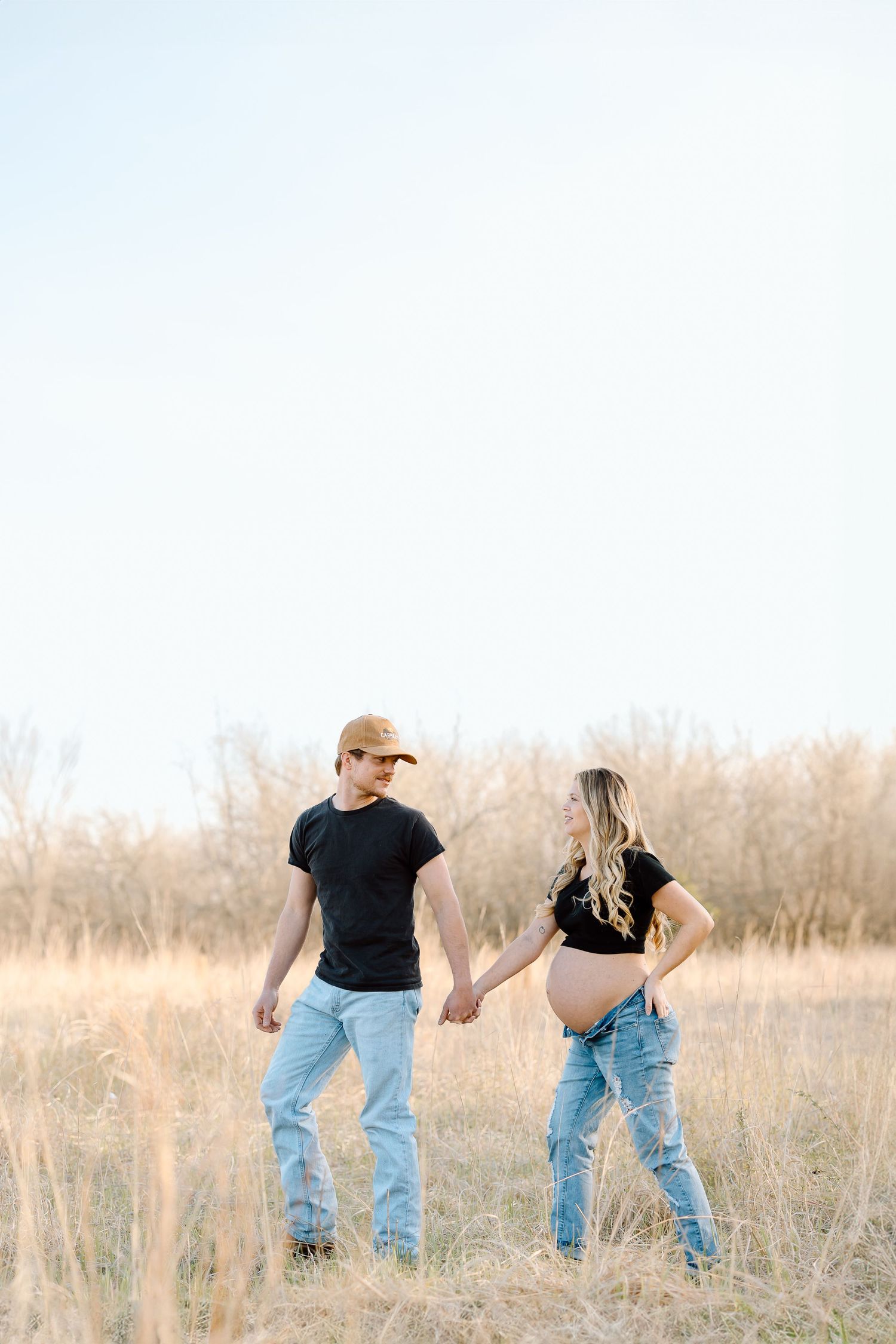 A couple in matching black shirts and jeans holds hands while walking through a golden field at sunset.