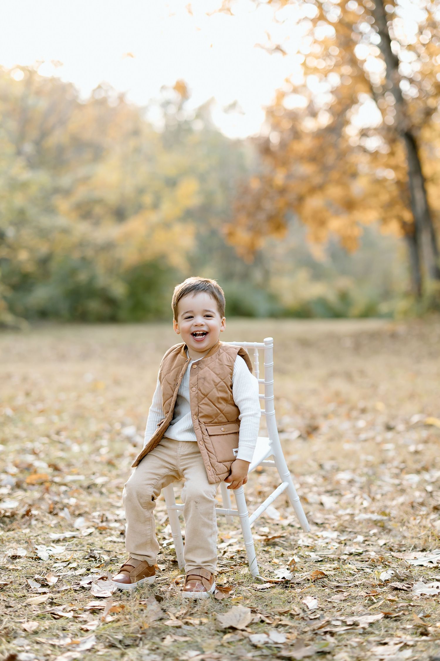 A child sits in a white wooden chair surrounded by fallen autumn leaves on a sunny fall day.