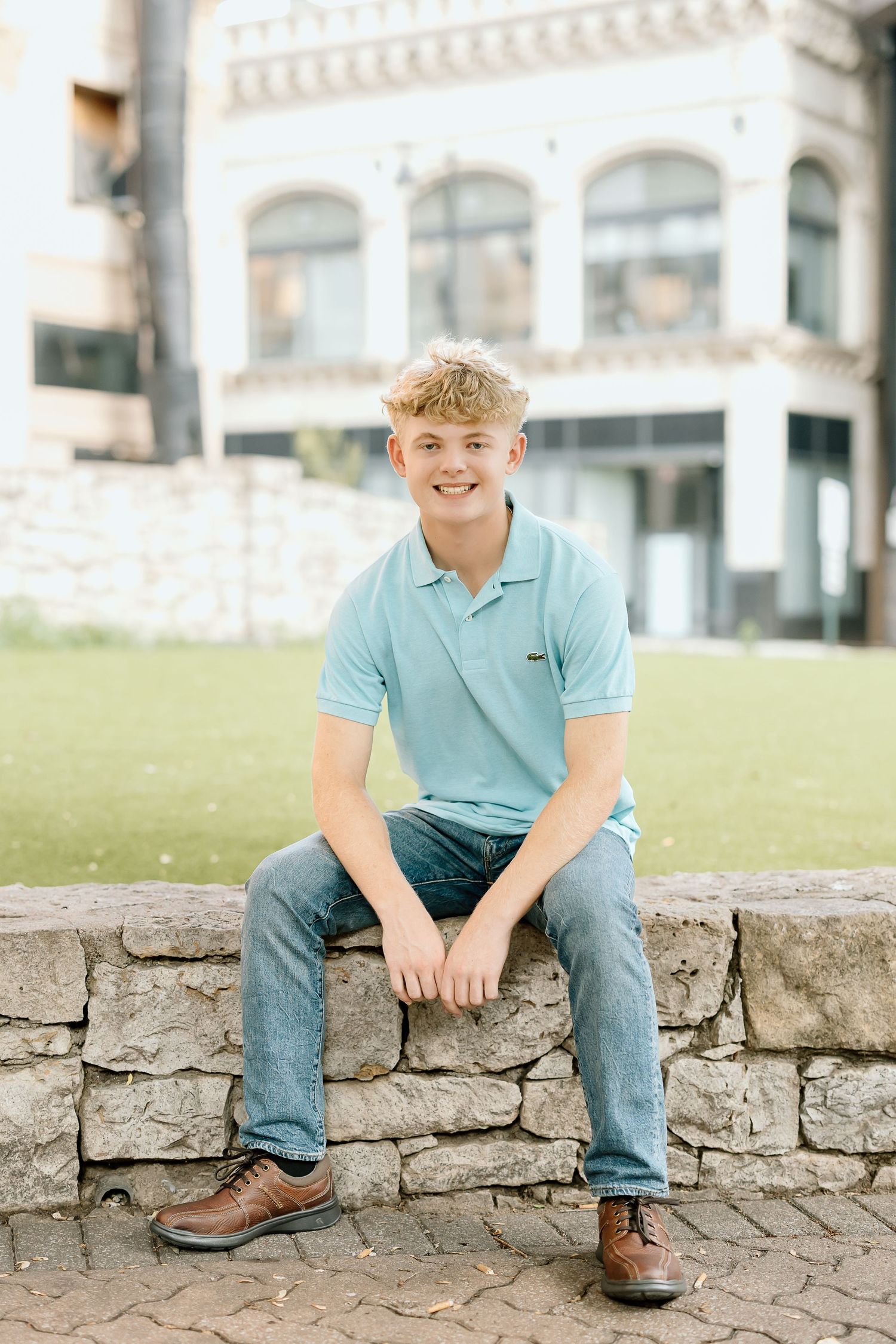 A person in a light blue polo shirt and jeans sits casually on a rustic stone wall outdoors on a sunny day.