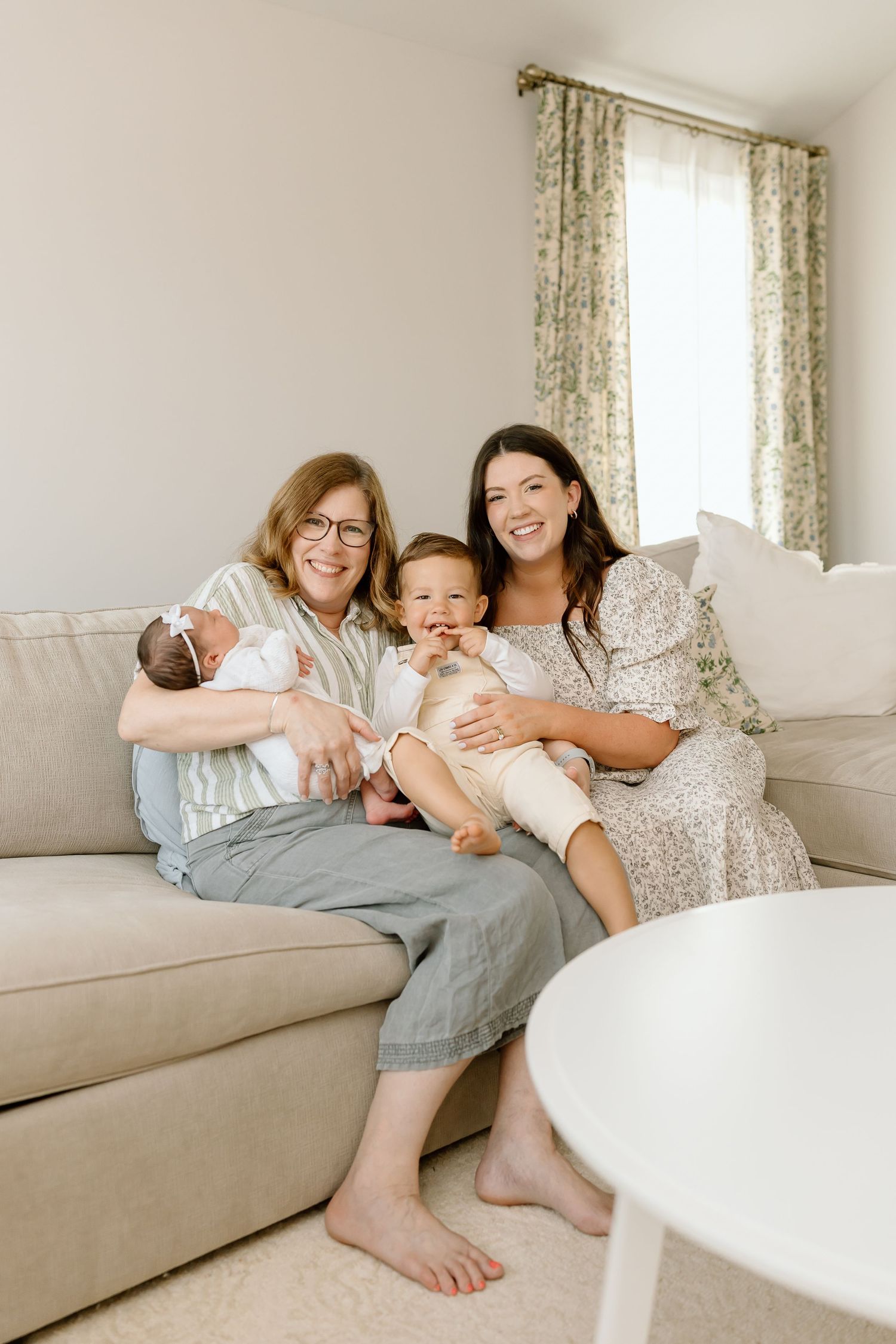 A family lounges together on a beige couch in a bright living room with floral curtains and natural lighting.