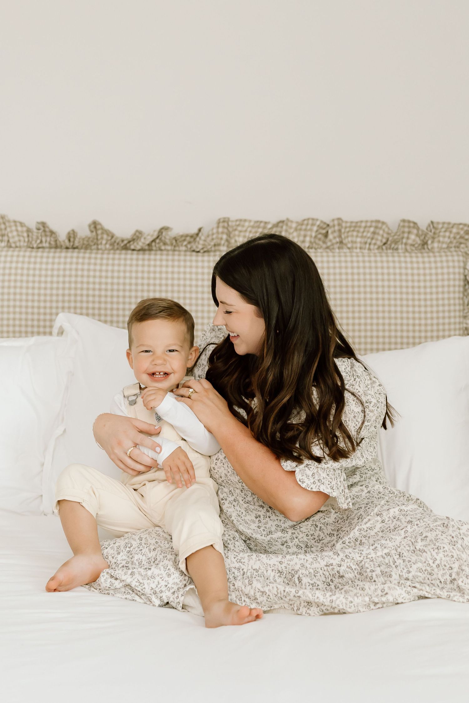 A mother and baby share a tender moment while laying together on white bedding with a gray floral blanket.