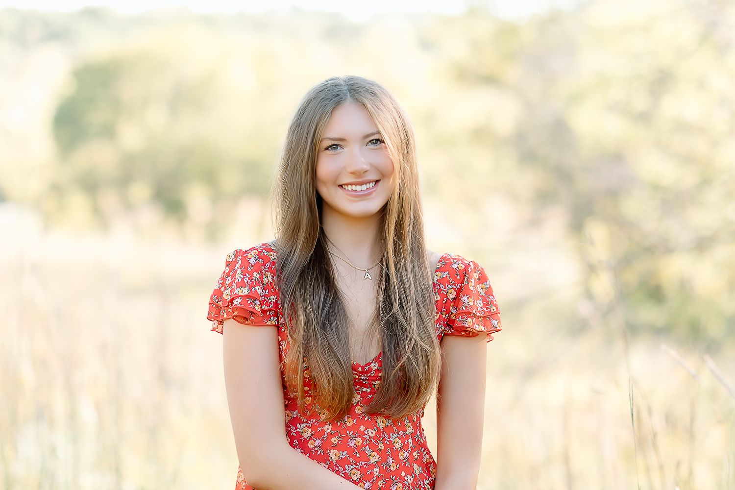 A person in a red floral dress sits outdoors in a sunlit field with tall grass and soft bokeh background.