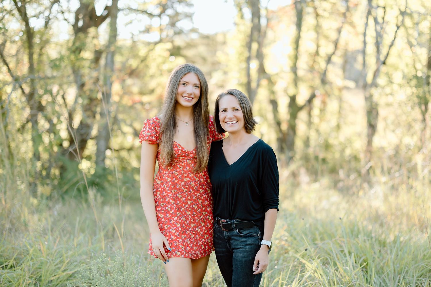 Two people stand together in a sunlit wooded area during golden hour.