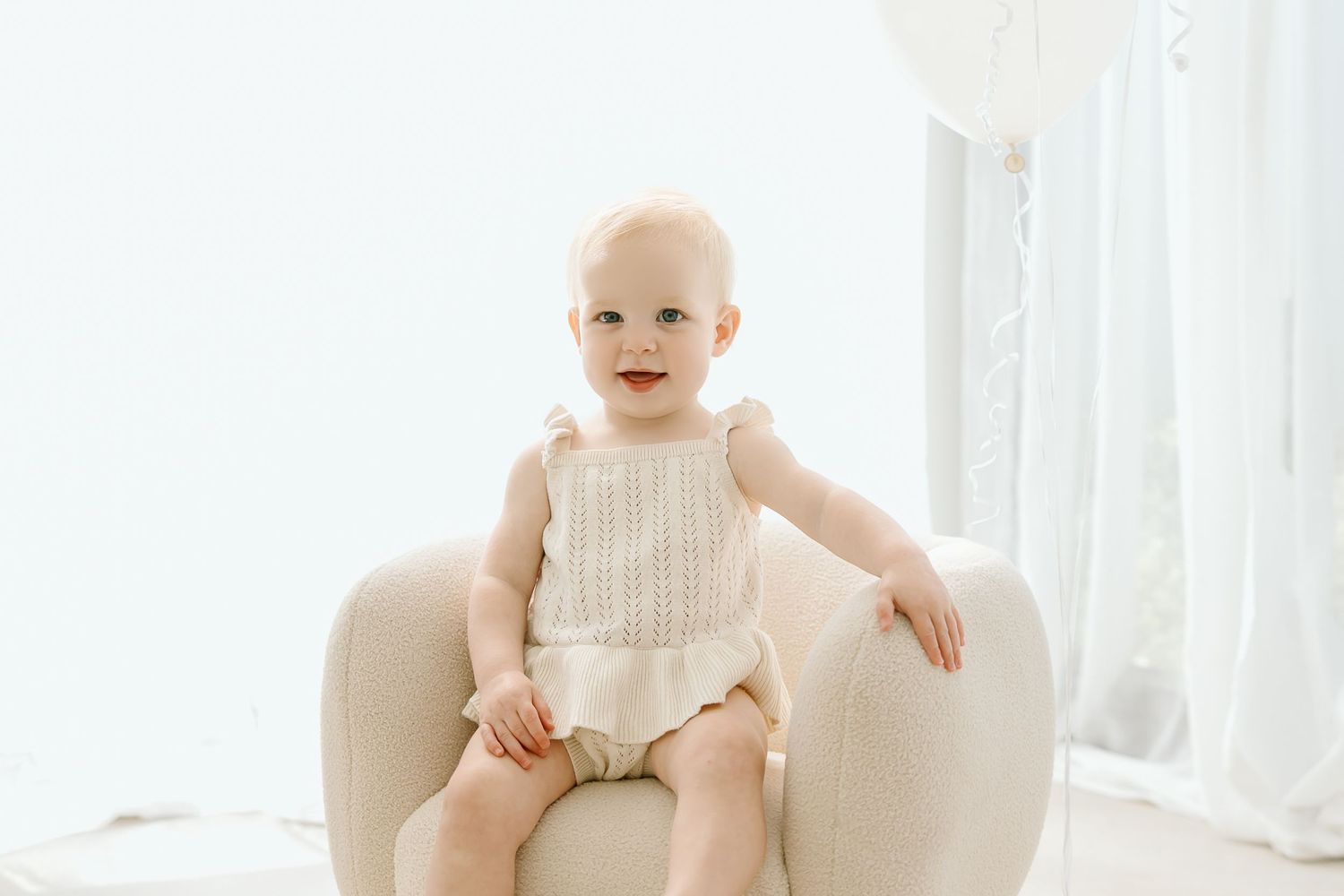 A baby wearing a white sleeveless dress sits smiling in a cream-colored armchair by sheer curtains.