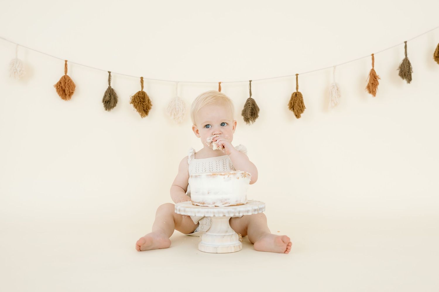 Small baby in white outfit explores birthday cake on cream background with autumn leaf garland.