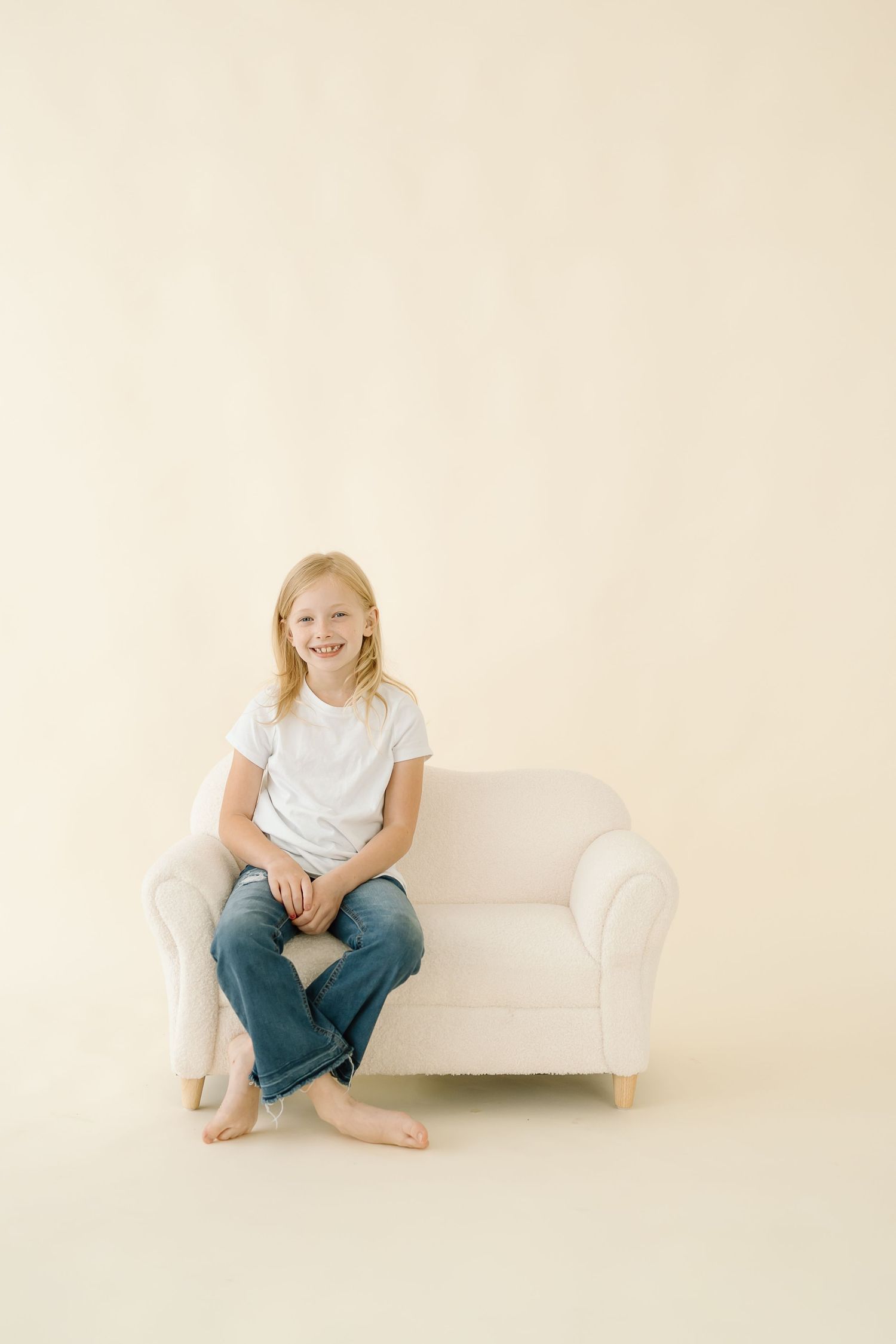A child sits cross-legged and barefoot on a white loveseat against a neutral beige background.