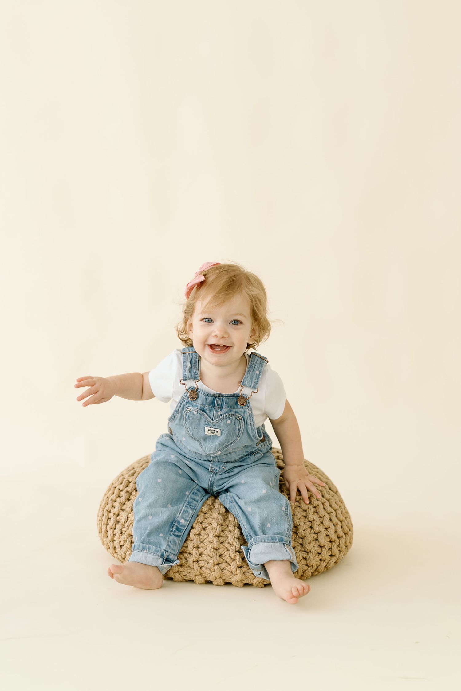 Baby in denim overalls sits and plays on a beige knitted pouf against cream background in progression photos.